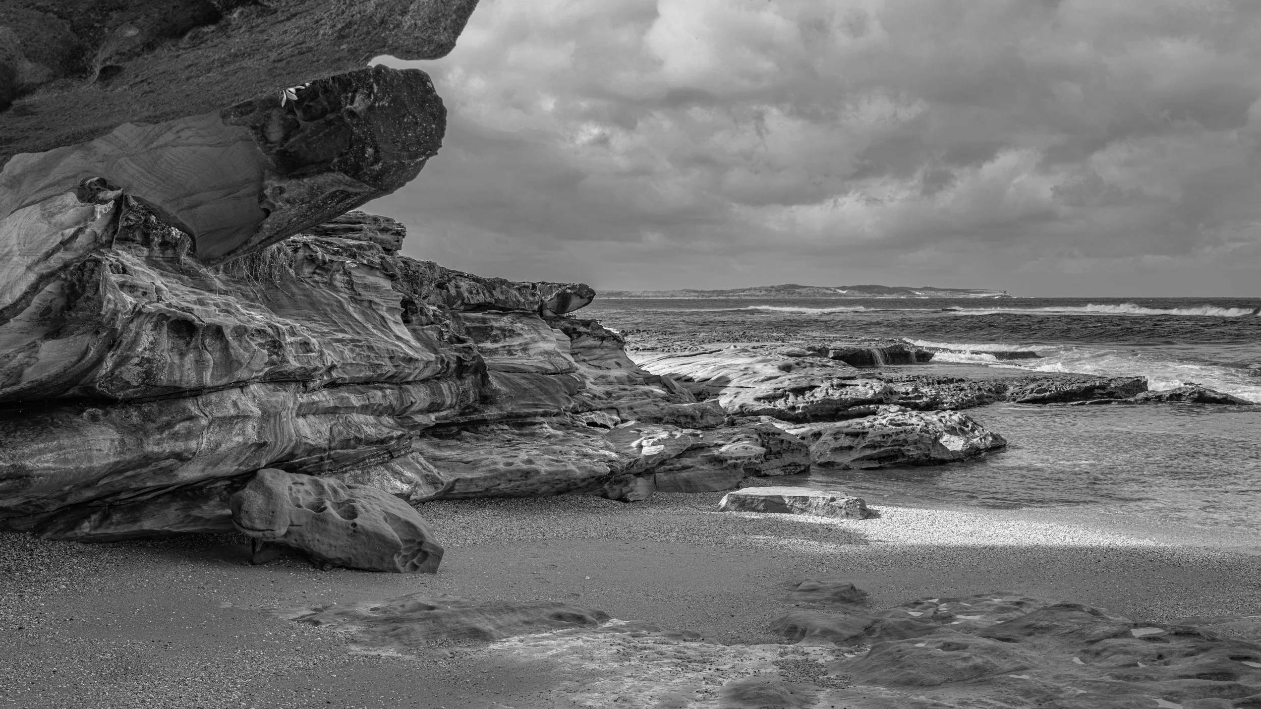 Black and white photo of a rocky beach with large layered rock formations on the left, extending into the water, with the ocean waves and cloudy sky in the background.