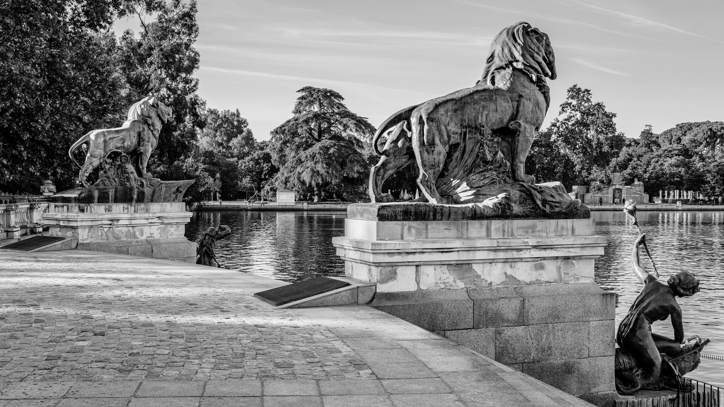 Black and white photo of two large lion sculptures on marble pedestals beside a water body, surrounded by trees and a paved walkway.