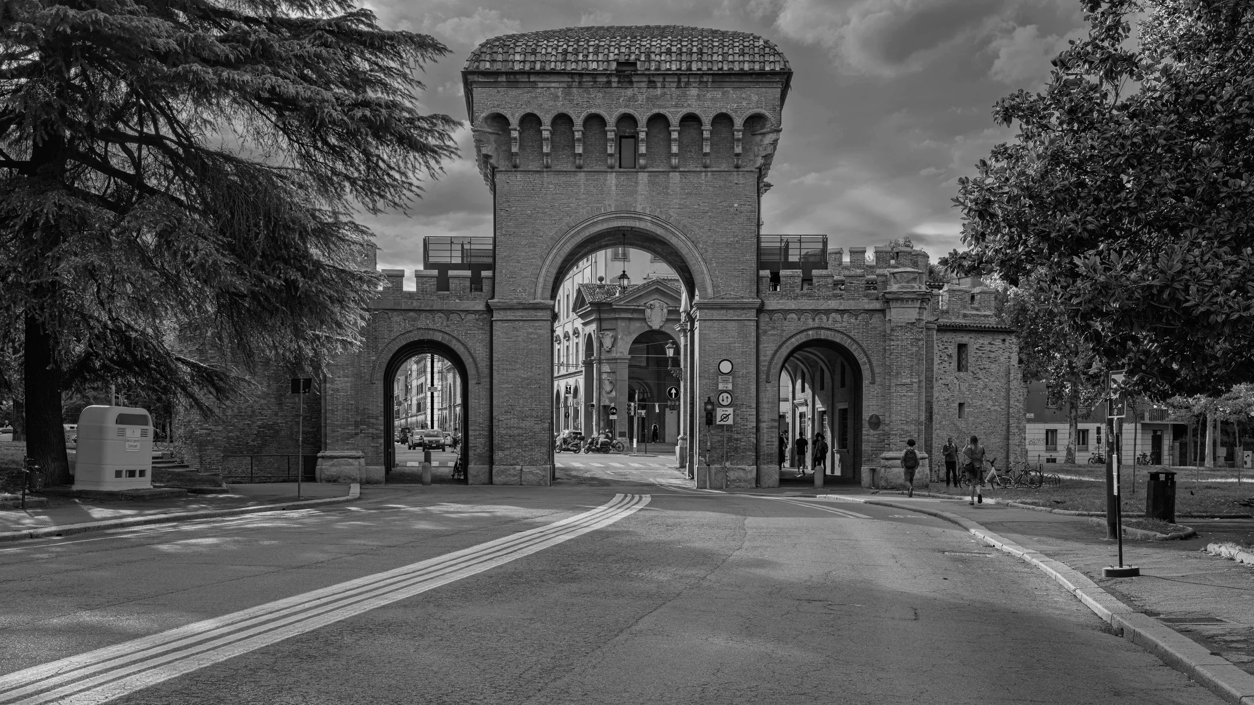A historic stone gate with an archway, flanked by trees, with people walking and cars passing underneath, and a cloudy sky in the background.