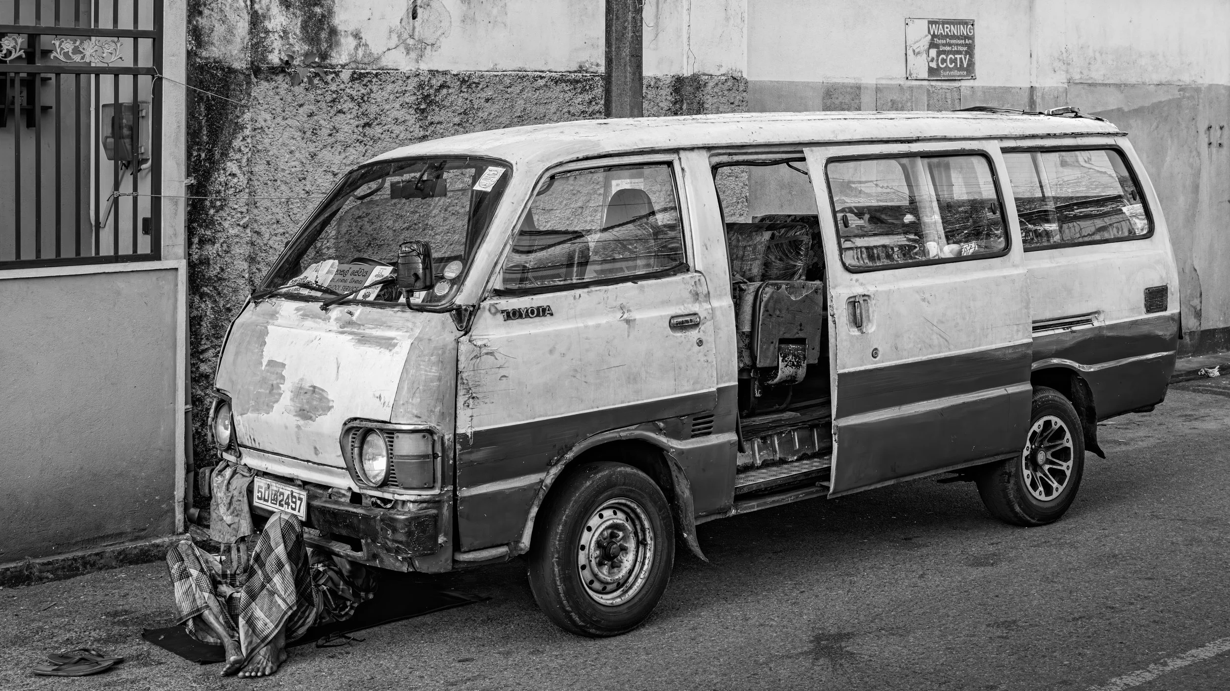 An old, dilapidated minivan parked on a street with debris and a cloth bag on the ground nearby. The background shows a wall with a warning sign about CCTV surveillance.