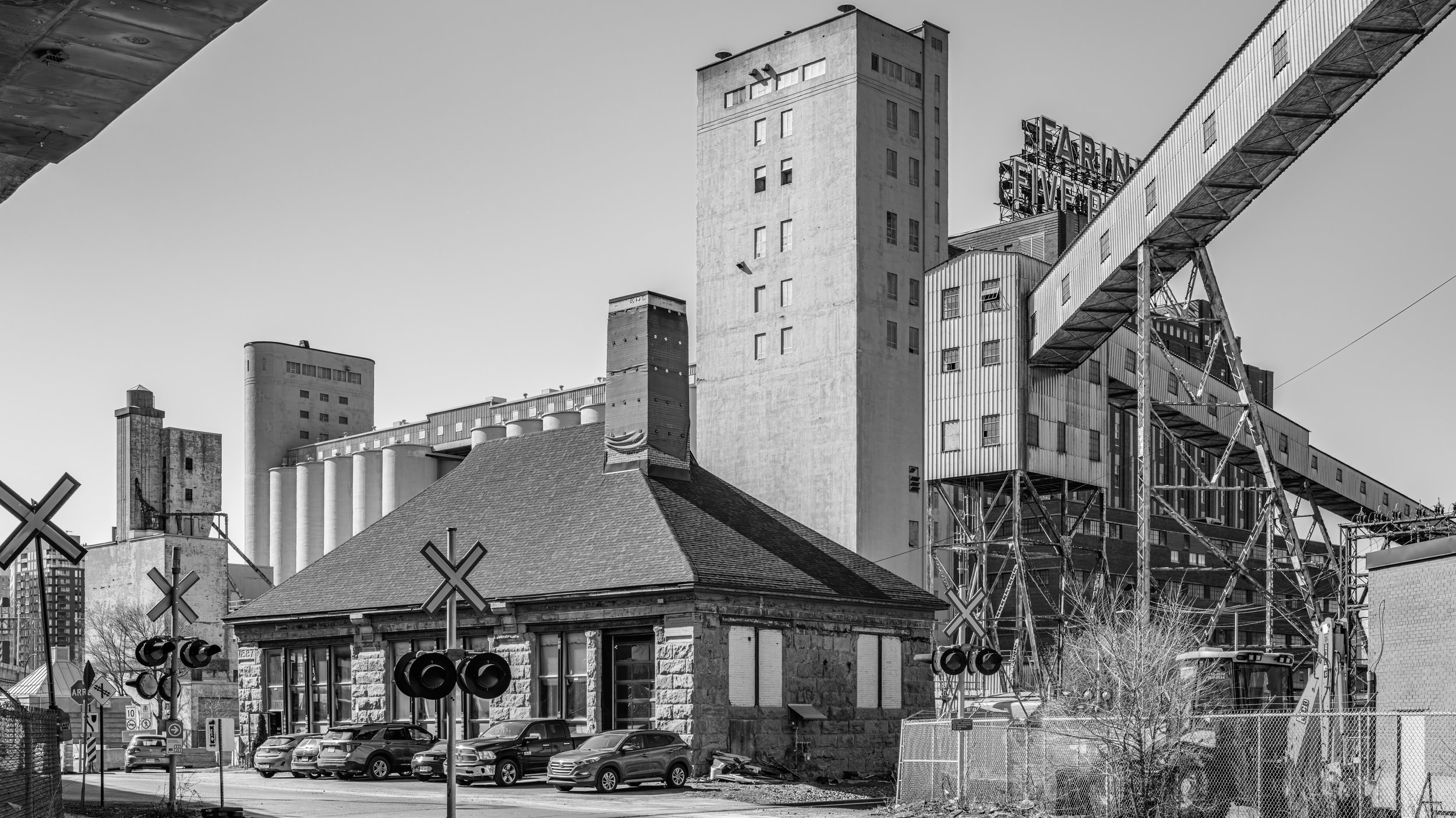 Black and white photo of urban buildings, including a large concrete grain elevator with a sign that says 'Farming Elevator,' next to a small house with a stone foundation and a tin roof, and a parking lot with cars.