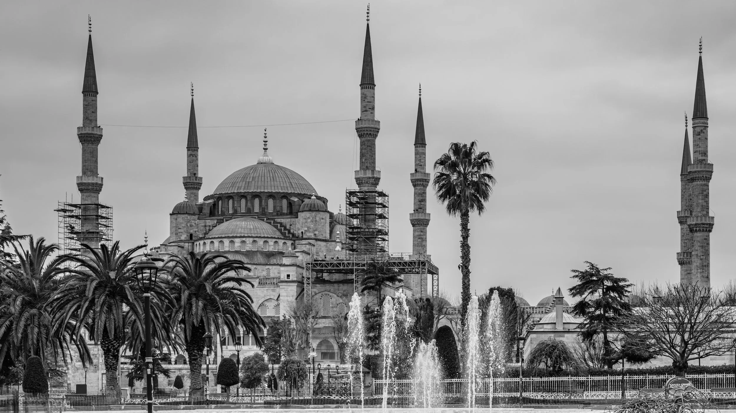 A black and white image of a mosque with six tall minarets, a large central dome, and smaller domes. In front, there are palm trees, water fountains, and street lamps.
