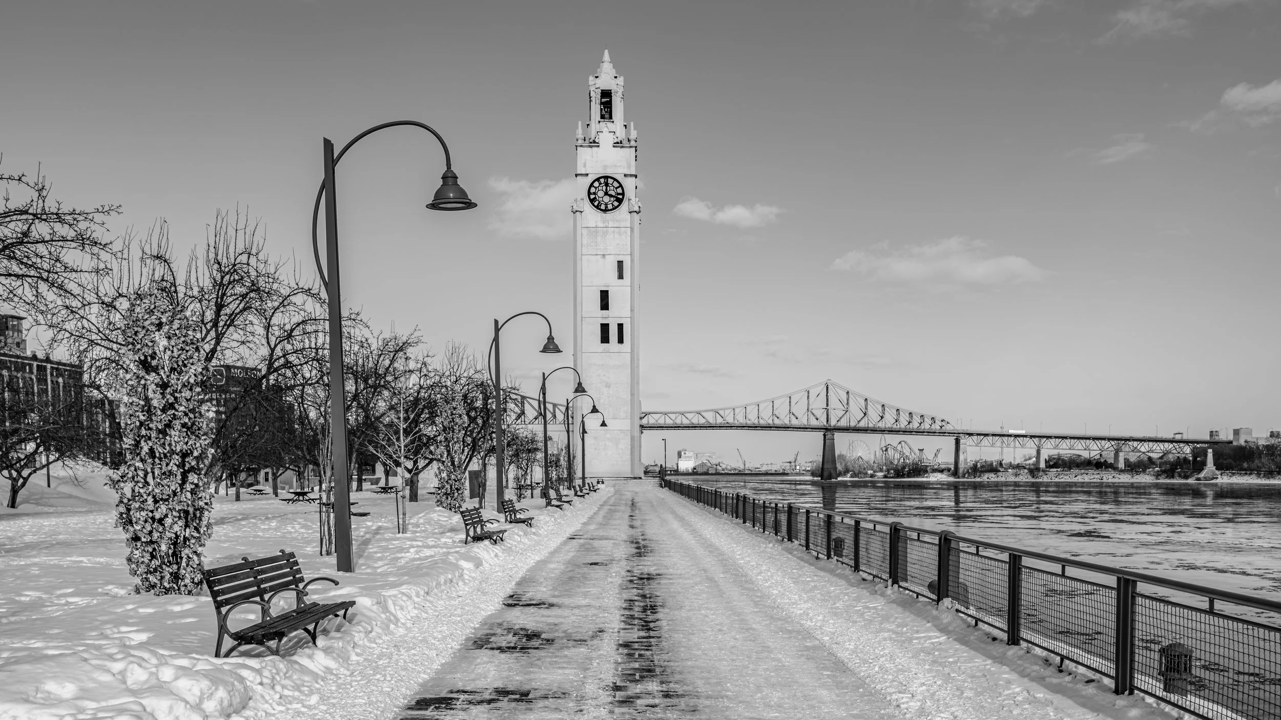 Black and white photo of a snowy waterfront park with benches, trees, and lampposts along a pathway. A tall clock tower and a bridge are visible in the background.