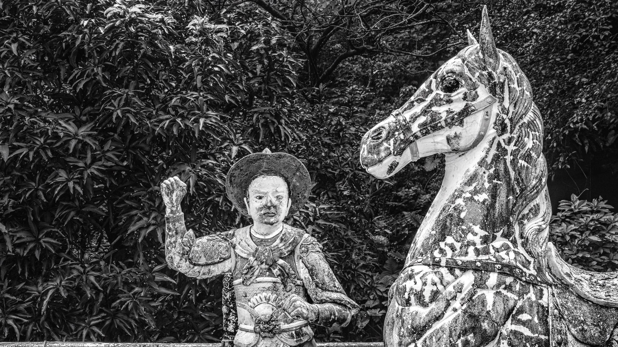 Black and white photo of stone statues of a young warrior with a raised fist and a unicorn with an elaborate mane, set against a background of dense foliage.
