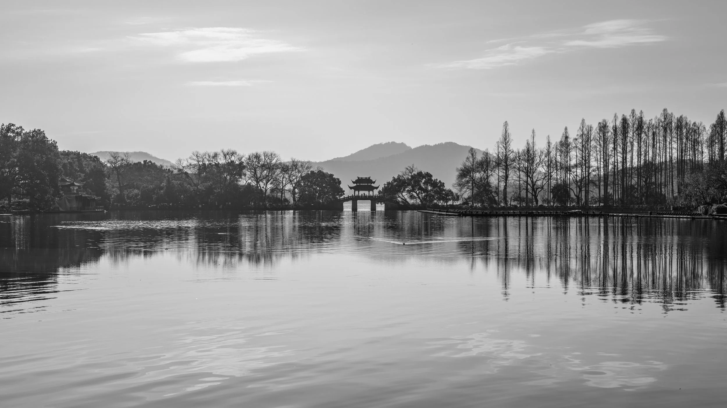 Black and white photo of a lake with a traditional Japanese-style pavilion on it; trees line the lake and mountains are in the background.
