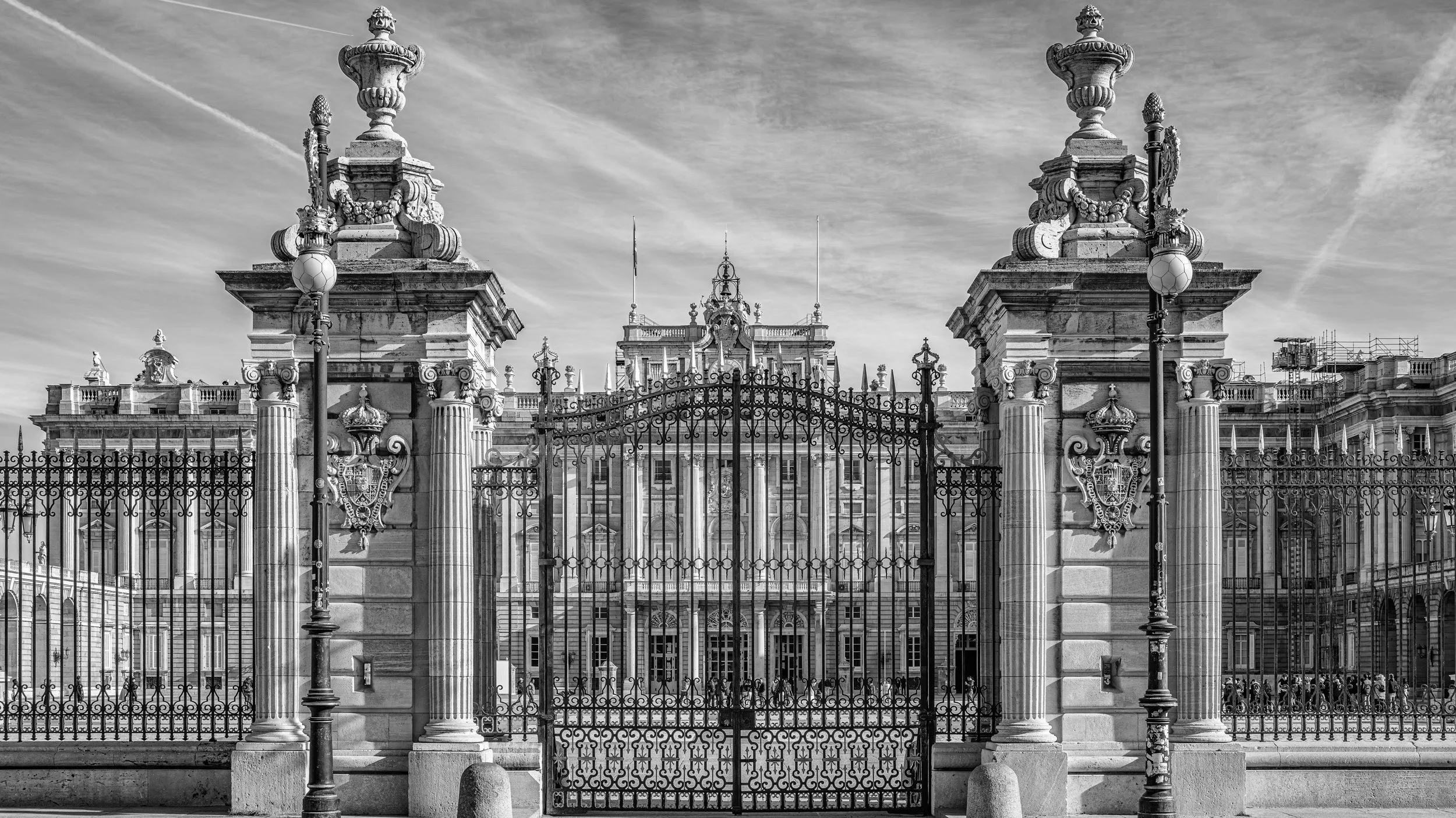Black and white photo of ornate palace facade behind a wrought iron gate with stone columns and decorative street lamps.