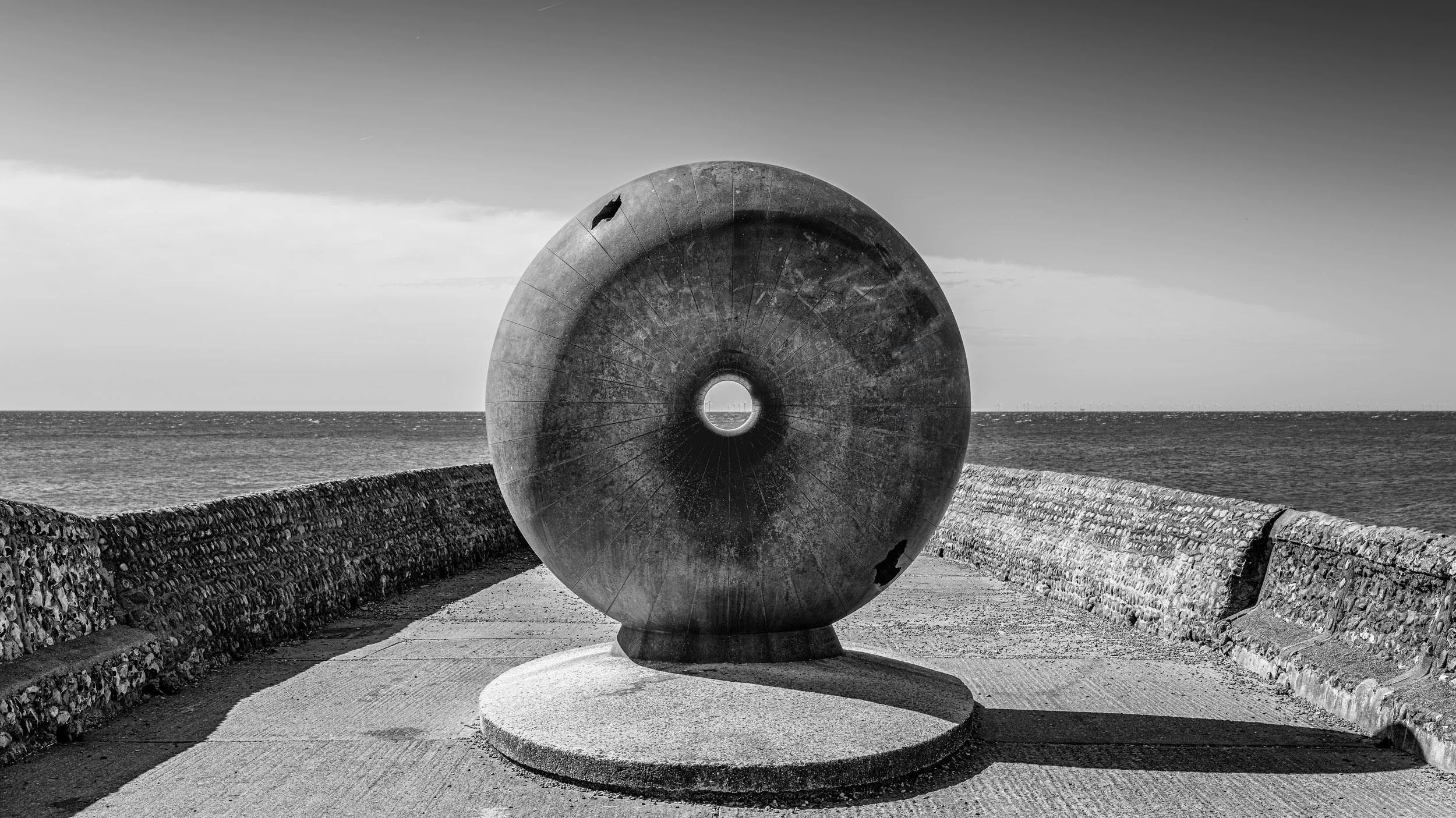 A large, circular, concrete sculpture with a central hole situated on a paved walkway near the ocean, bordered by stone walls on either side, with a cloudy sky in the background.