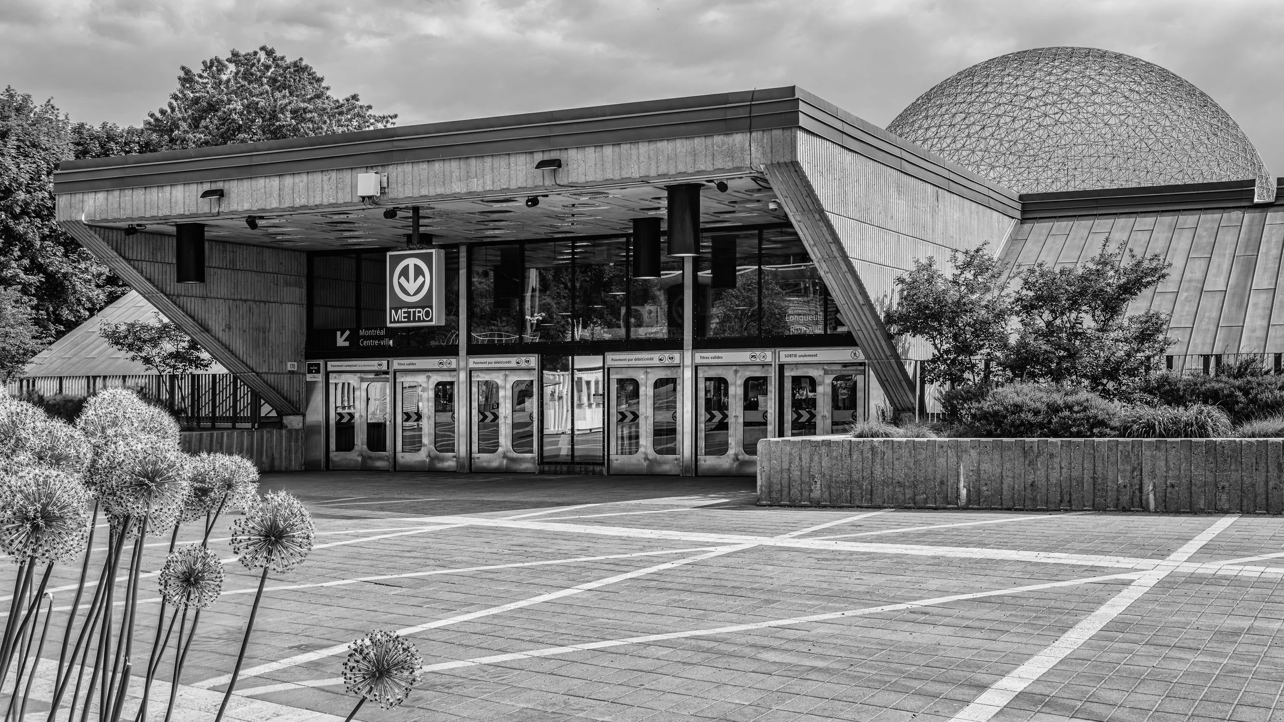 Black and white photo of a modern metro entrance building with a geodesic dome on the roof, surrounded by trees and plants, with an empty parking lot in the foreground. Montreal, Quebec, Canada.