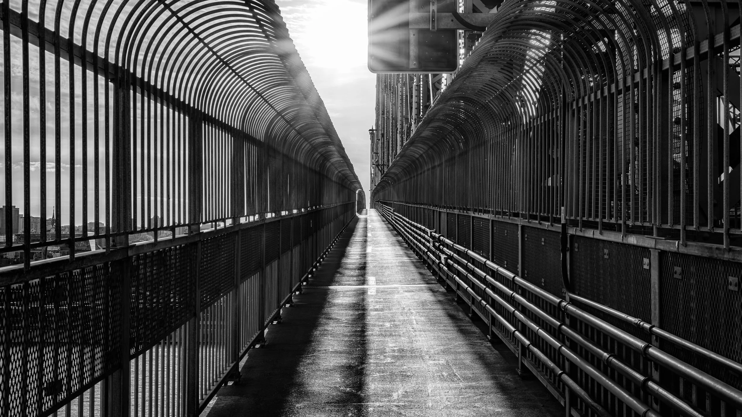 Black and white photograph of a high pedestrian bridge with metal railings and barriers, stretching into the distance with city buildings visible below.