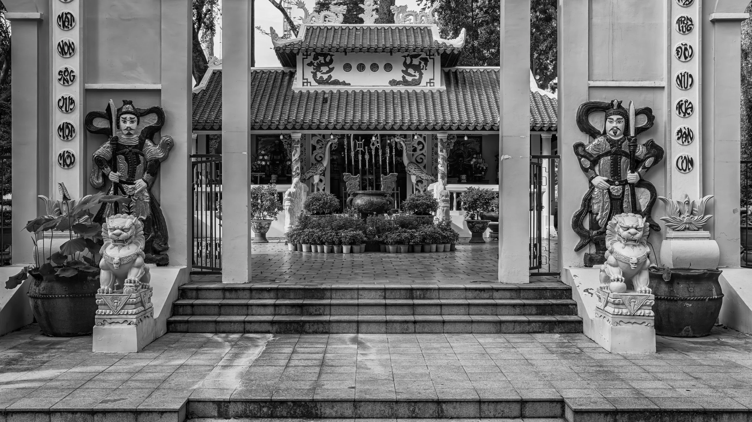 Black and white photo of a traditional Asian temple entrance with statues of warriors holding swords, statues of lions, and potted plants, with steps leading up to the temple.