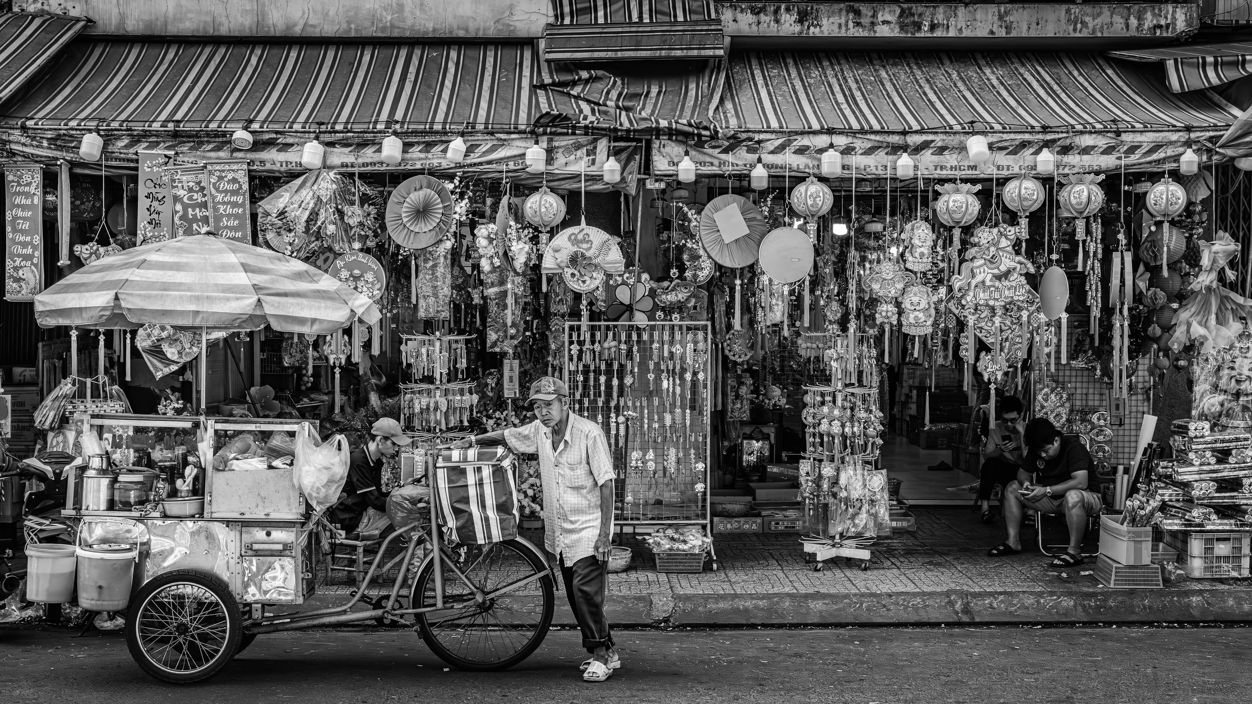 Street market stall with hanging lanterns, fans, and souvenirs, with two men and two women in front, some sitting and one walking, in black and white.