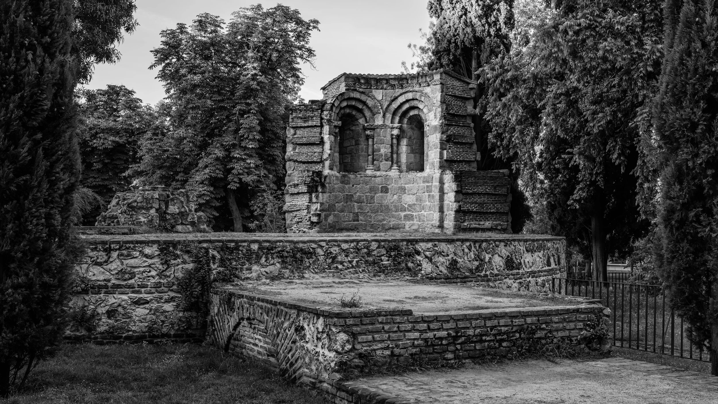 Ancient stone ruins with arches, surrounded by trees and overgrown foliage, in black and white.