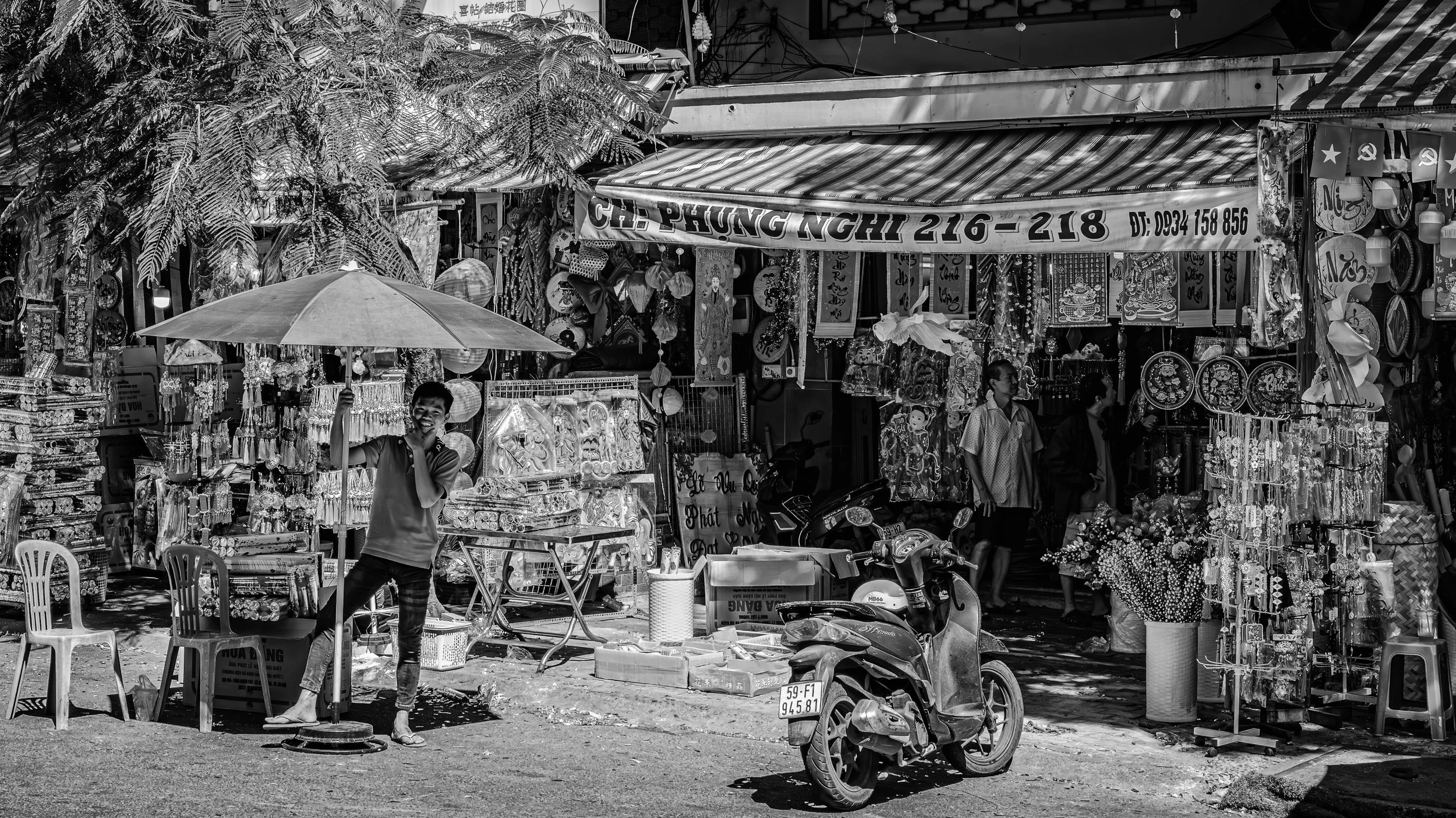 Street market scene with stalls selling jewelry and decorations, people browsing, a man holding an umbrella, motorcycles parked in front, and a tree providing shade.