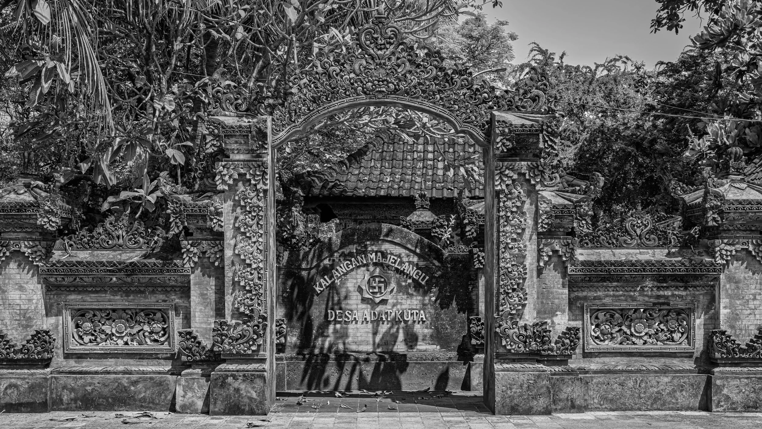 Ornately carved traditional Balinese entrance gate with intricate floral and swirl designs, surrounded by dense tropical foliage, with a visible partly tiled roof behind it, and a sign that reads 'Kalangan Majelangu Desa Adat Kuta' with a Balinese sw