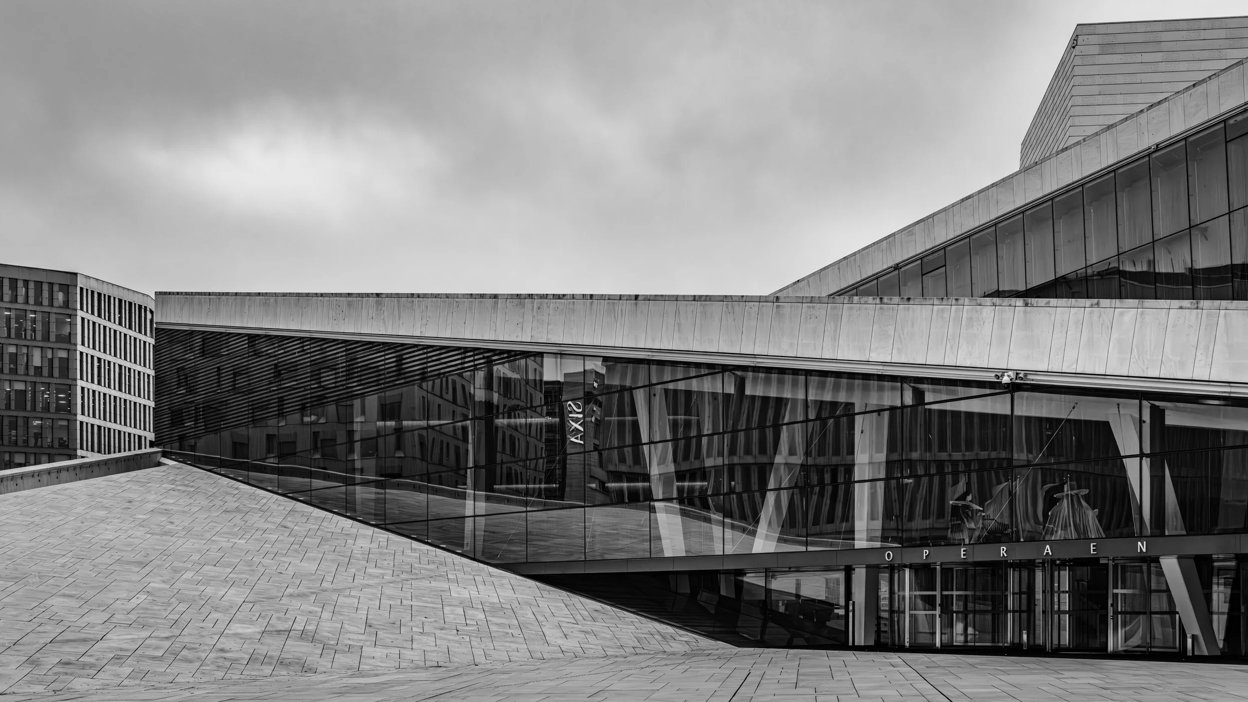 Modern building with large glass windows, curved roof, and the word 'OPERA' visible near entrance, set against cloudy sky.