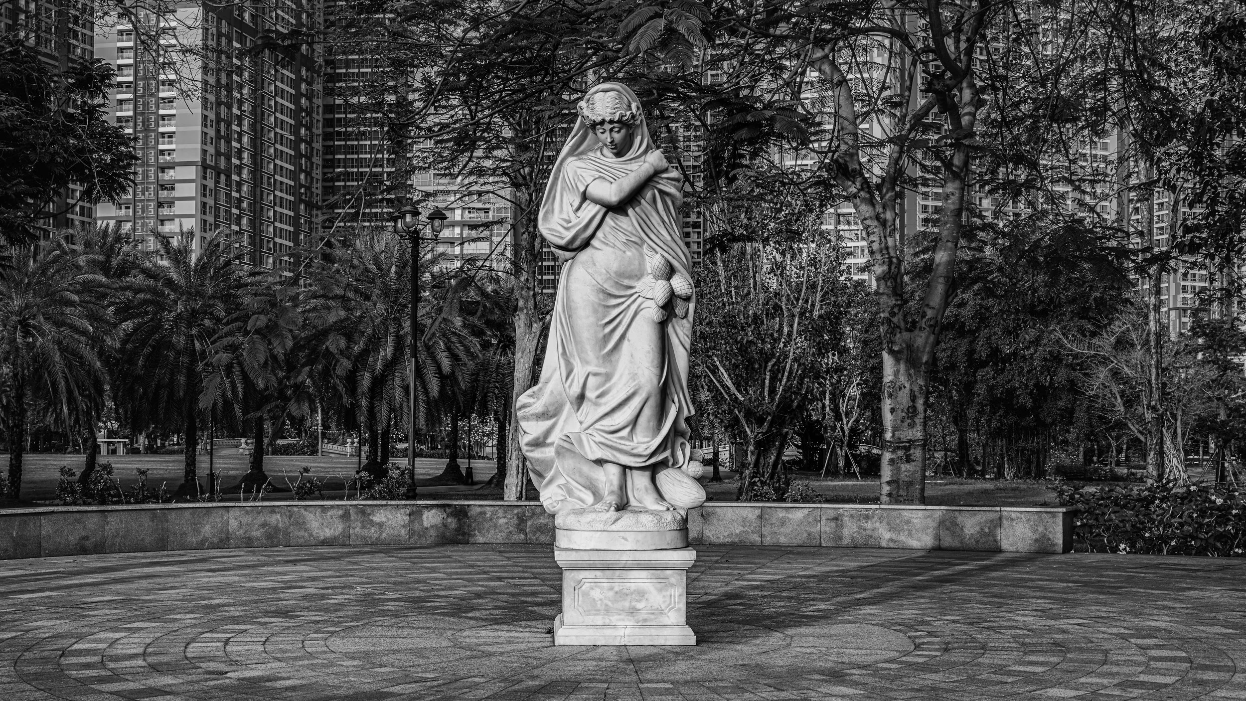A white marble statue of a woman in historical attire standing in a park, with tall modern apartment buildings and trees in the background.