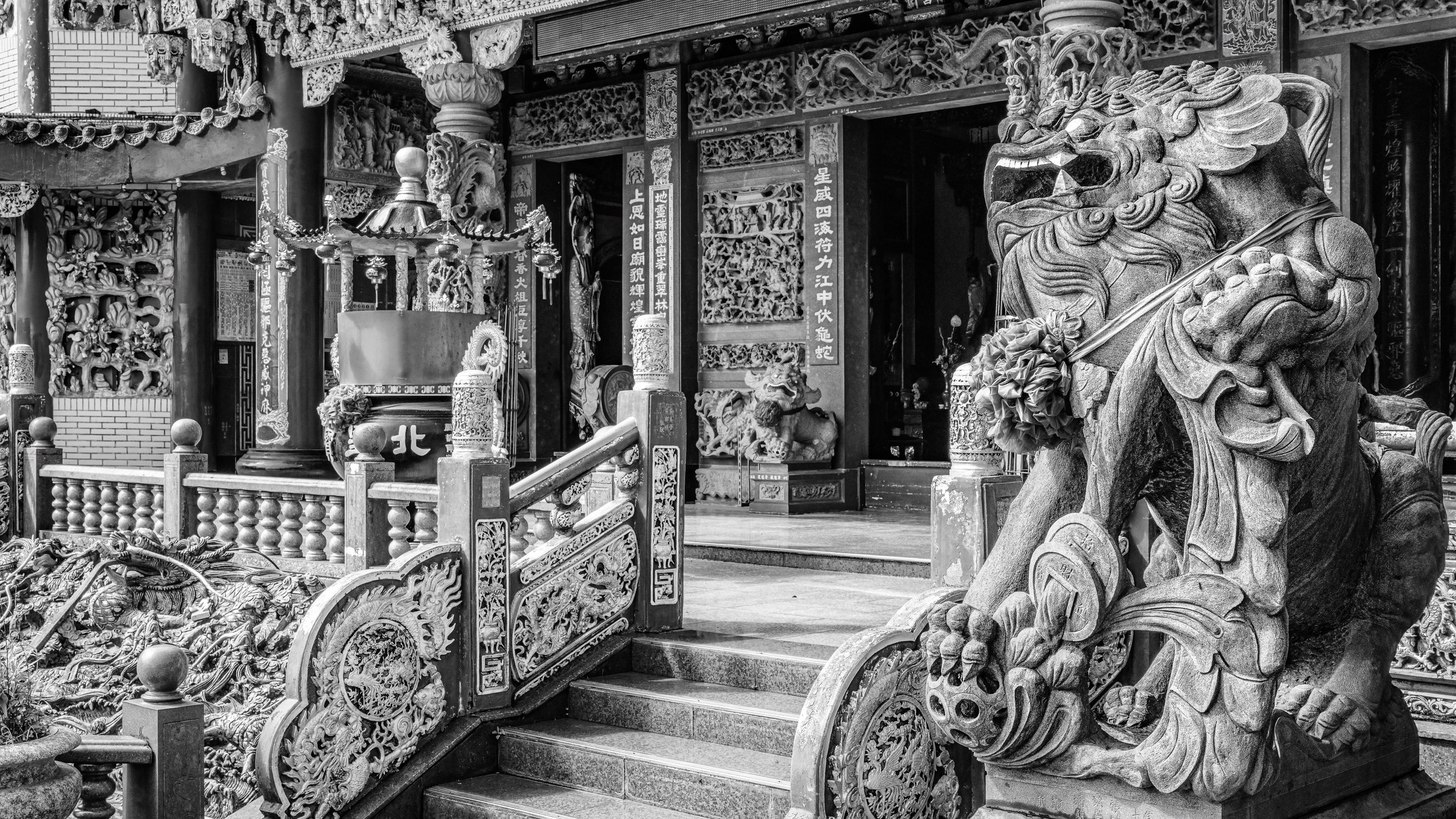 Black and white photo of ornate traditional Chinese temple architecture with detailed carved woodwork, stone lion statue at the entrance, and decorative elements.