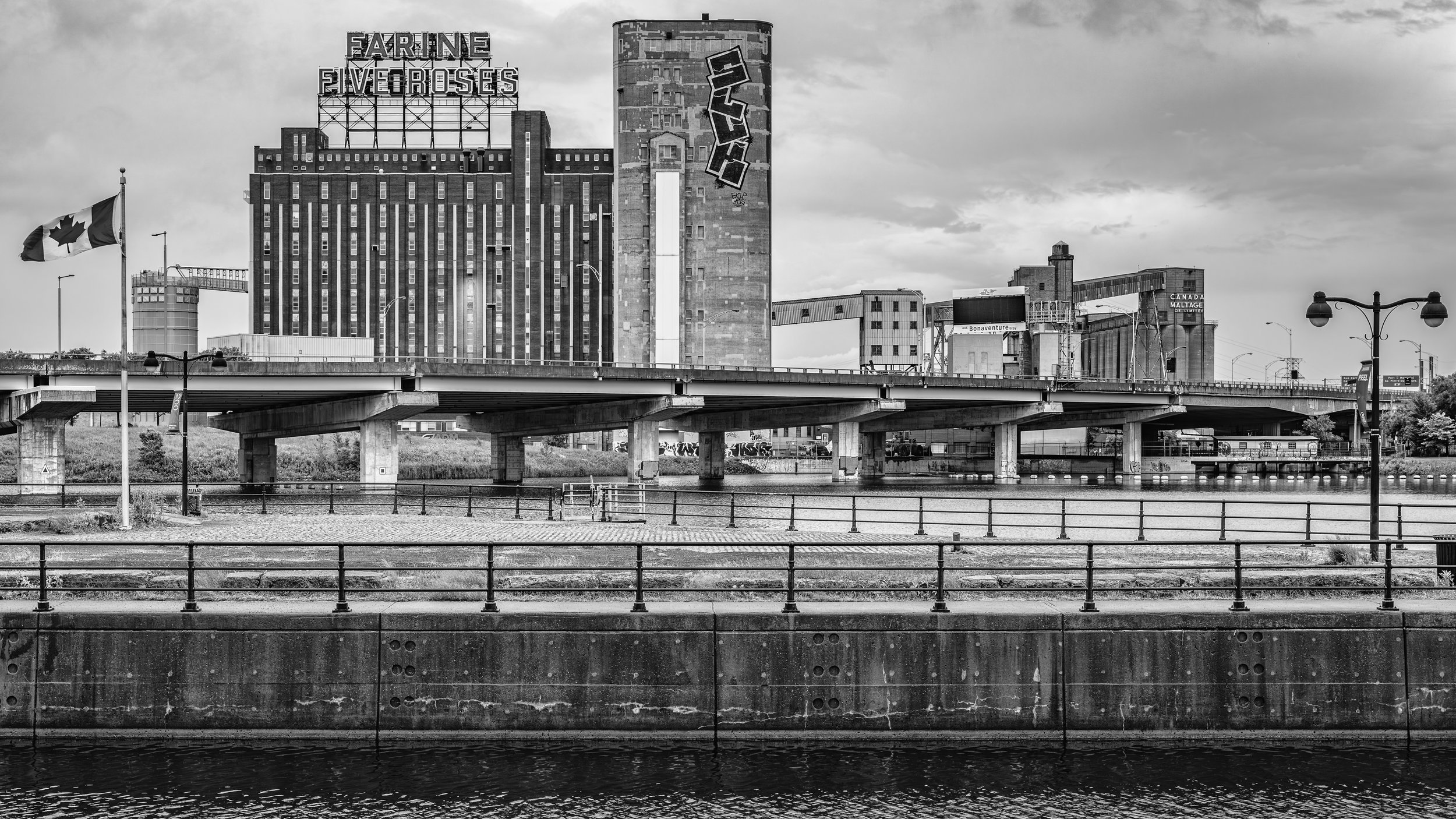 Black and white photo of urban cityscape with high-rise buildings, a bridge over a river, street lamps, and a Canadian flag.