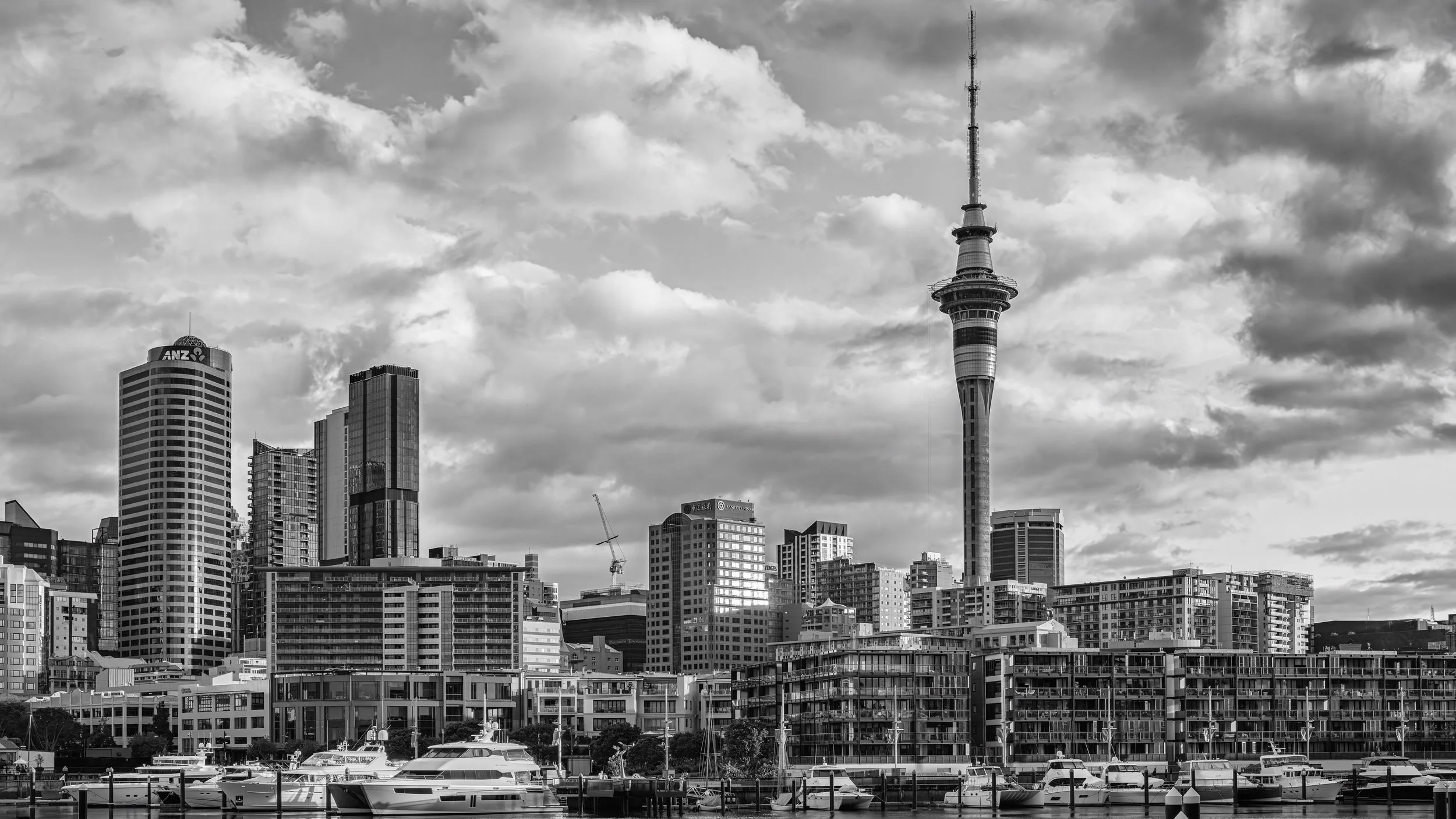 Black and white photo of a city skyline with various high-rise buildings, including a prominent tall tower with a circular observation deck, and boats docked in a marina in the foreground.
