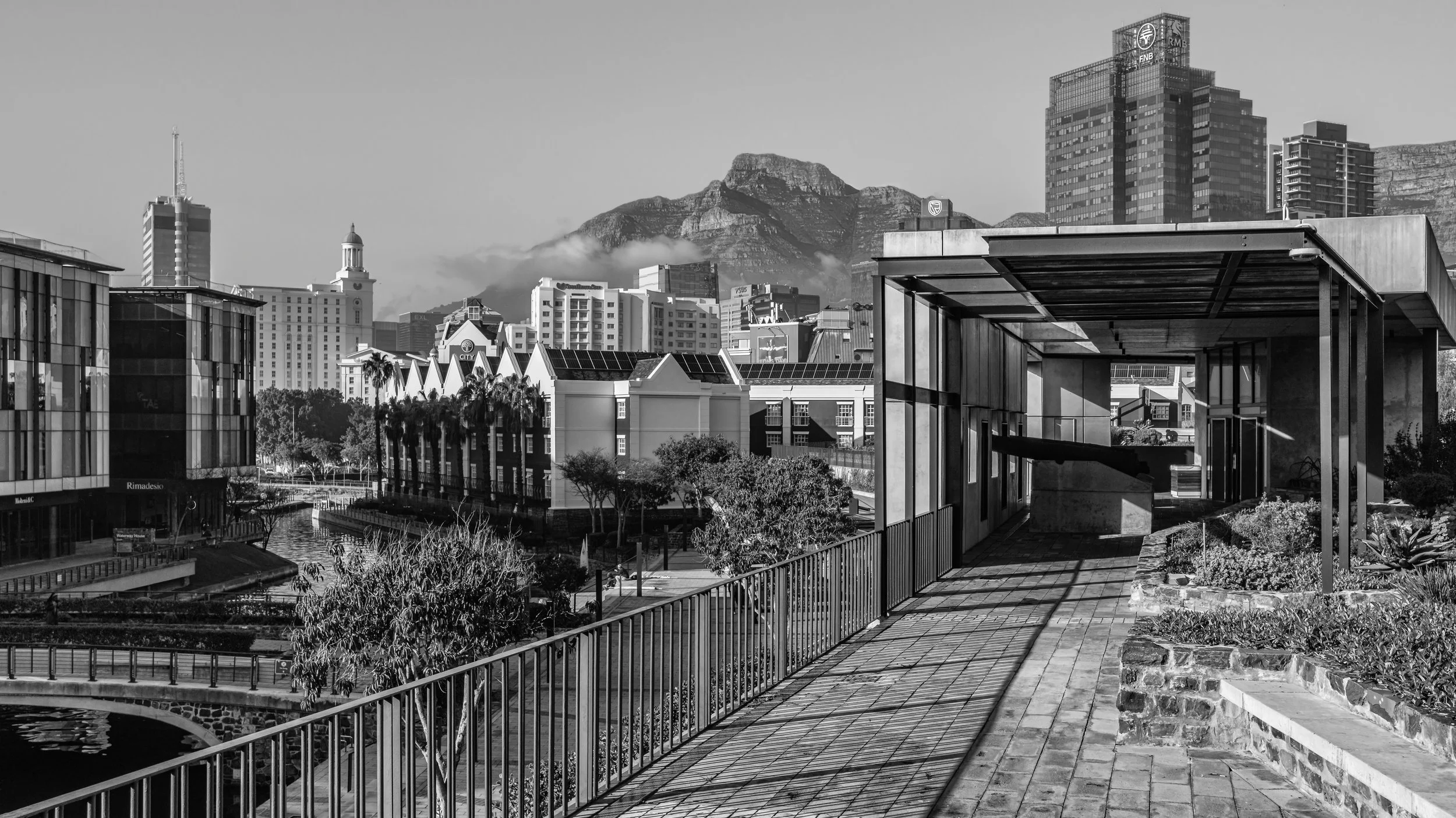 A cityscape with modern buildings, a tall skyscraper, and mountains in the background, viewed from a rooftop terrace with railings and a shaded seating area. Cape Town, South Africa.
