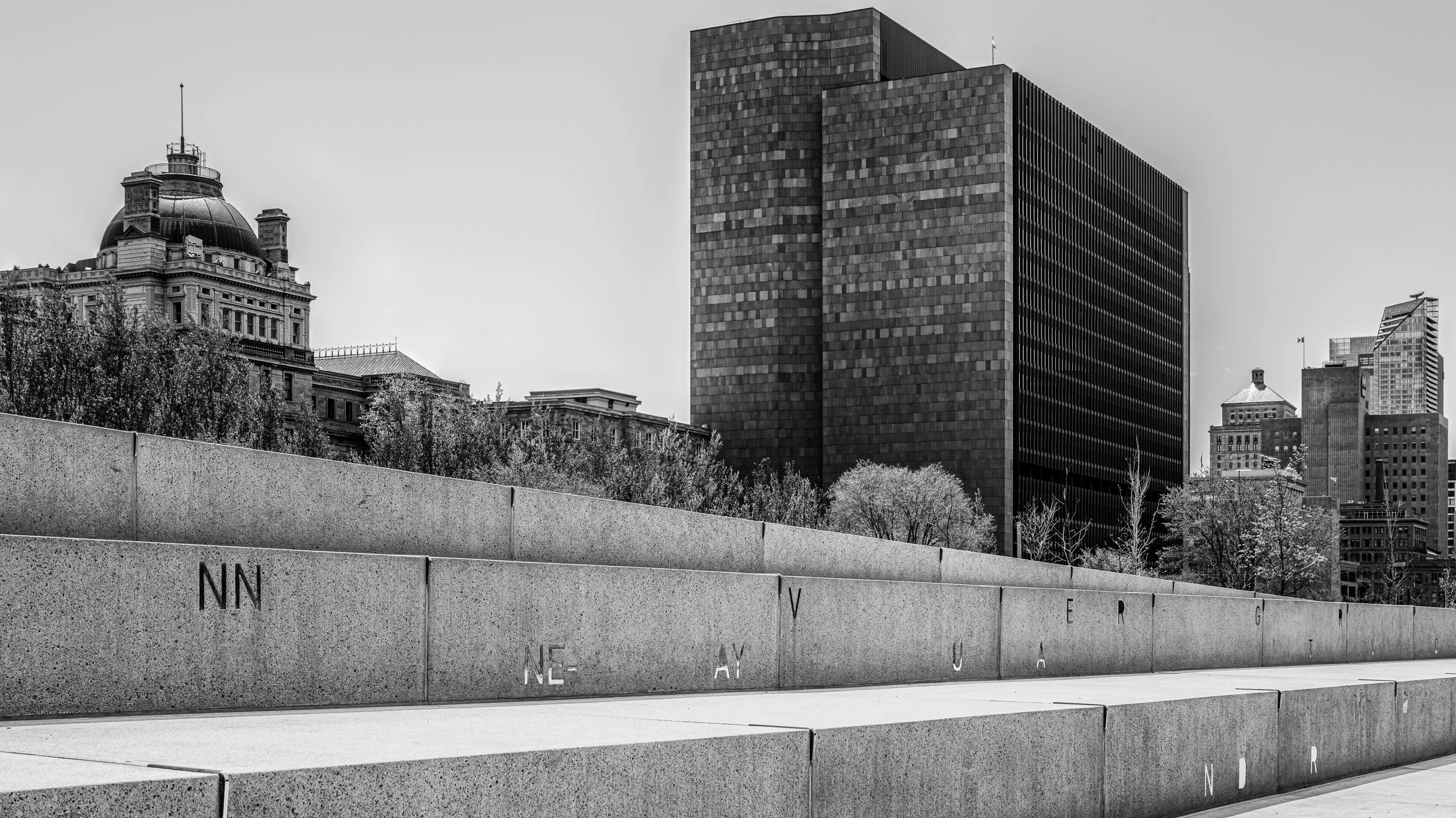 Black and white photo of a cityscape with modern skyscrapers and historic buildings in the background, viewed from a low angle with a stone wall in the foreground.