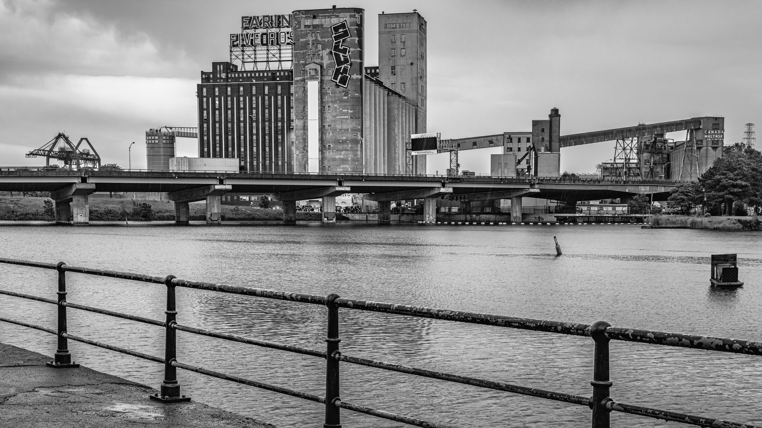 A black and white photo of an industrial cityscape viewed from across a body of water, with factories, silos, cranes, and buildings, including a high-rise with graffiti, under an overcast sky.