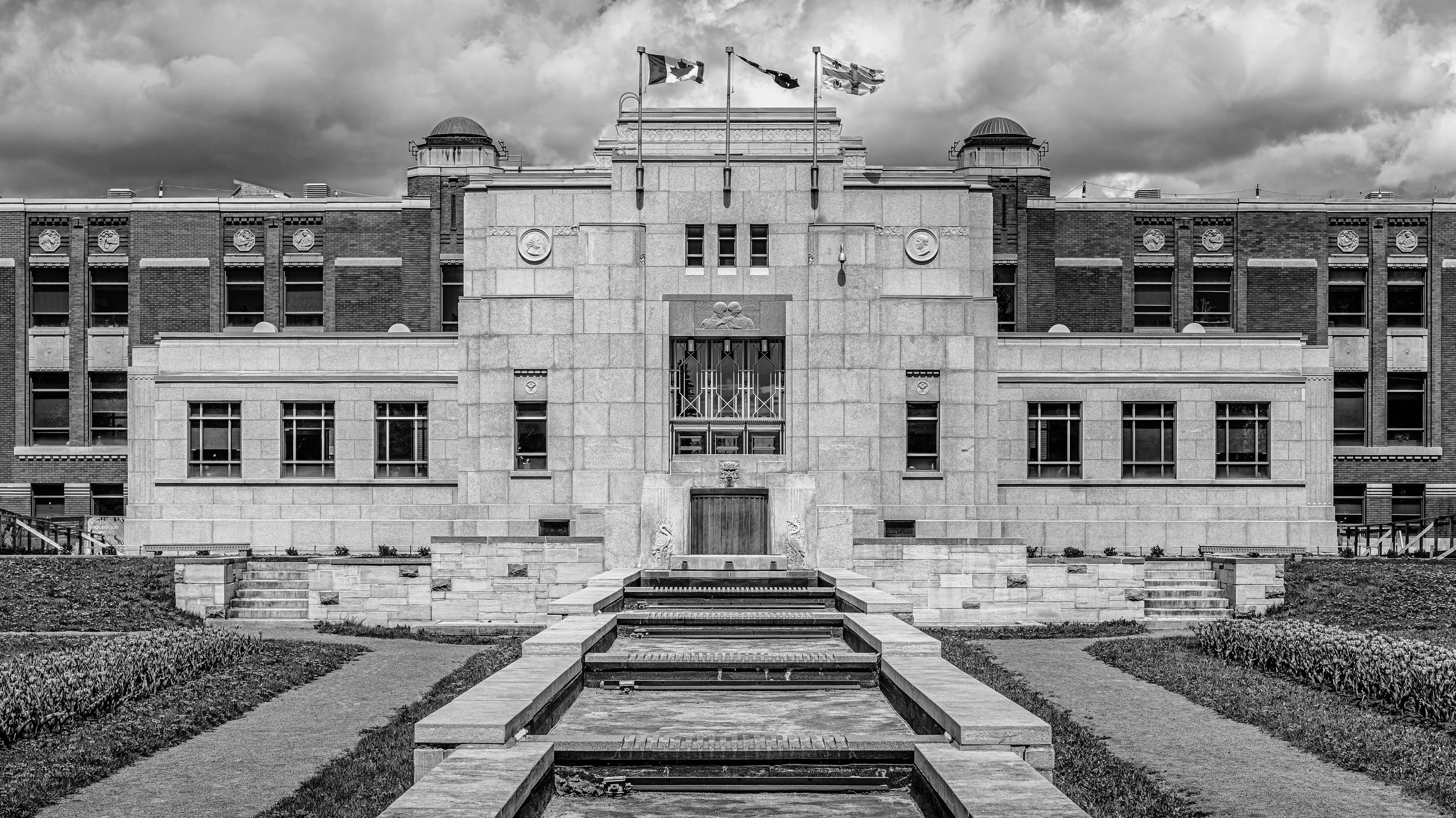 A historic government building with flags on top, surrounded by a garden with a pathway and water feature in front, under cloudy skies.