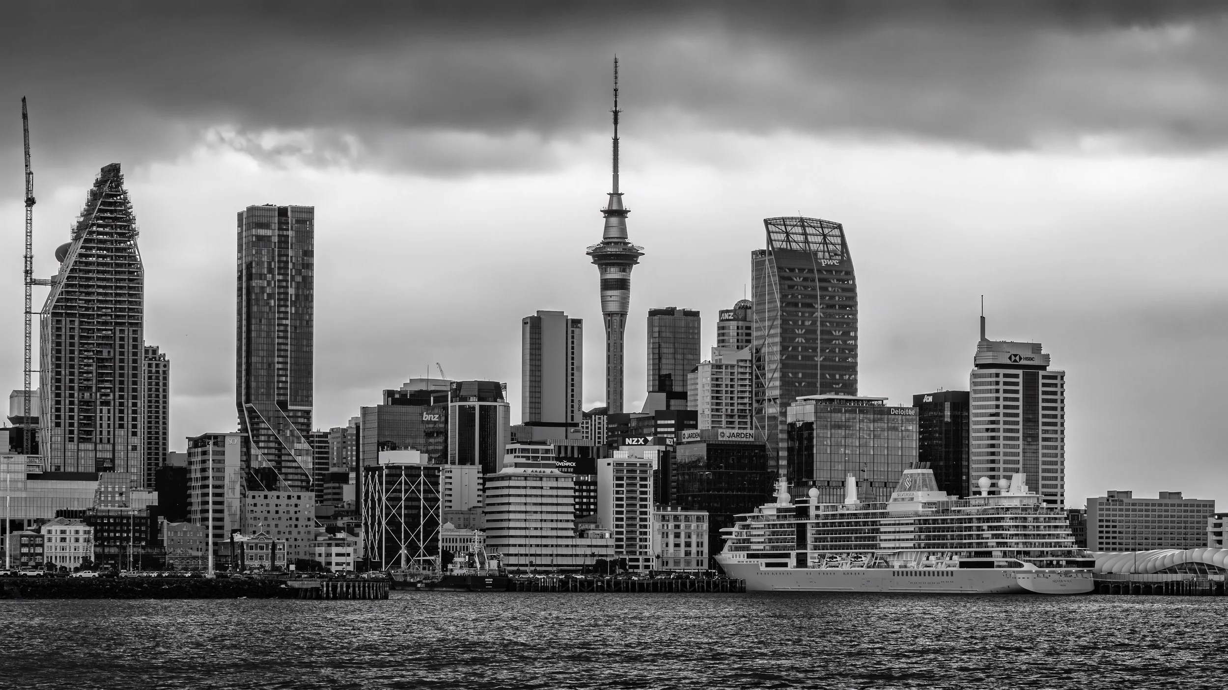 Black and white photo of a city skyline with various skyscrapers and a tall tower, viewed across a body of water.