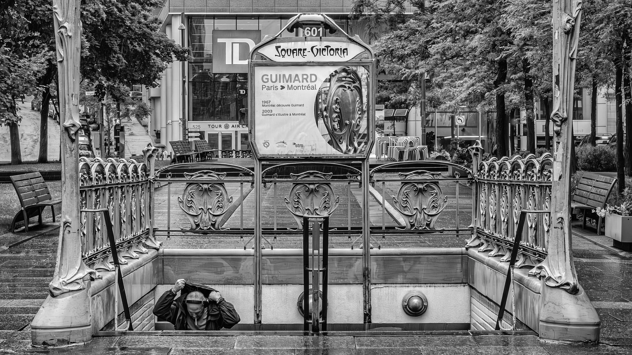 Black and white photo of an underground metro entrance with ornate metal railings, a sign for Square-Victoria station, and a billboard advertising Guimard bridges, with trees and buildings in the background.
