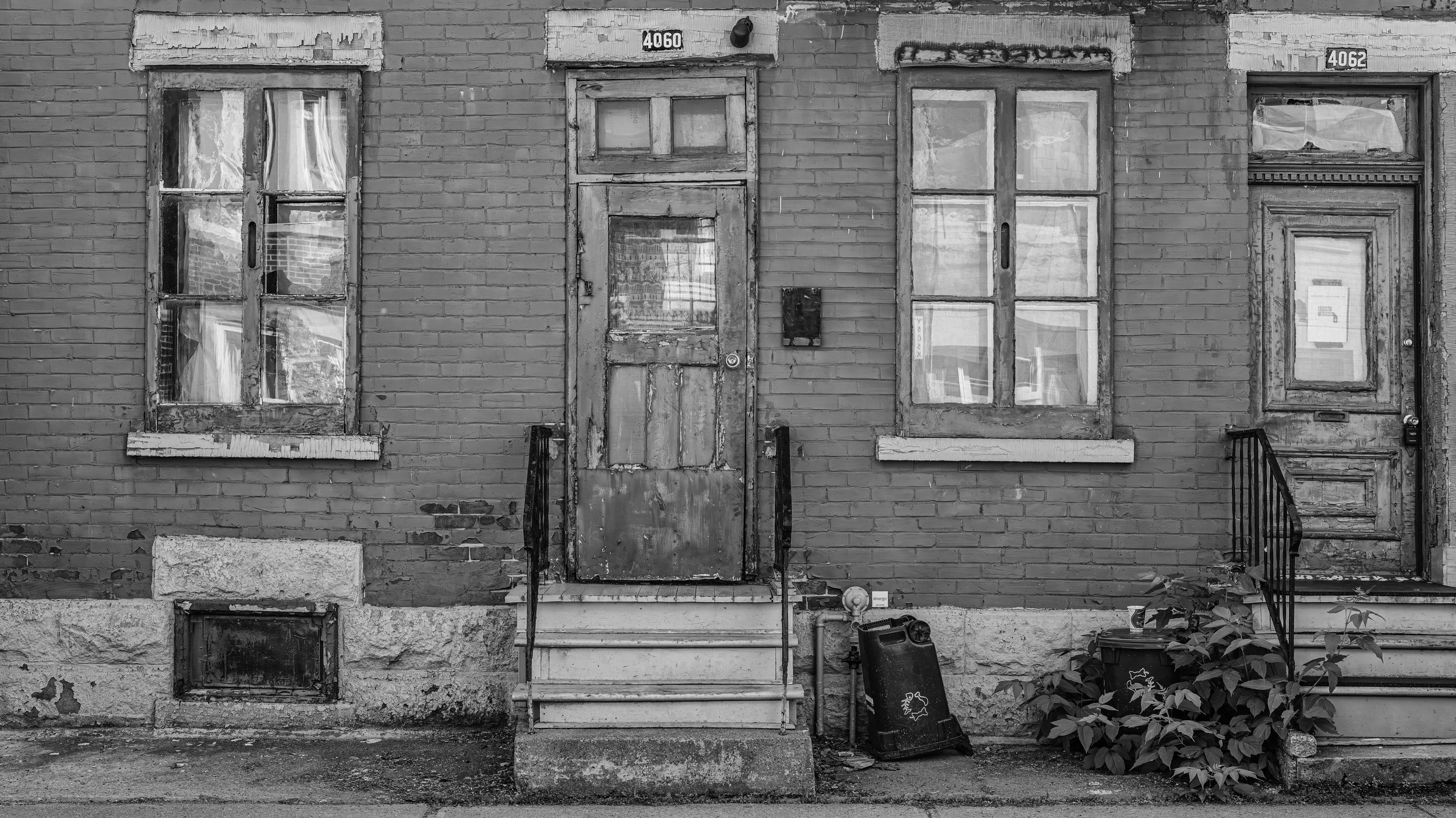 Black and white photo of an old brick house facade with worn-out windows and doors, metal stairs, and some plants near the bottom right.