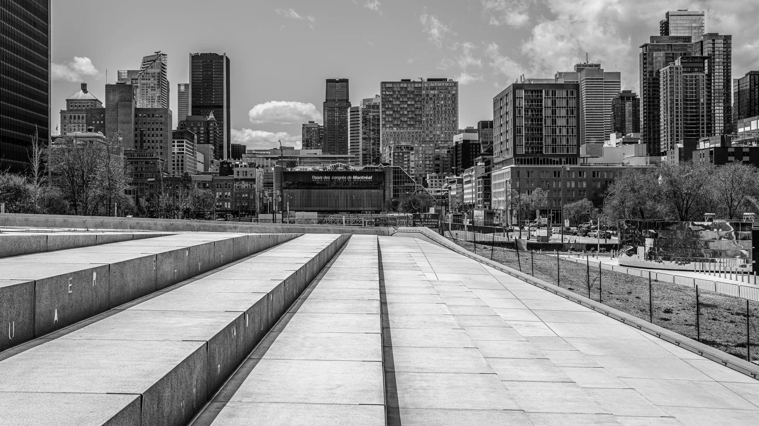 Black and white photo of a city skyline with tall buildings and a park or plaza with steps and a walking area in the foreground.