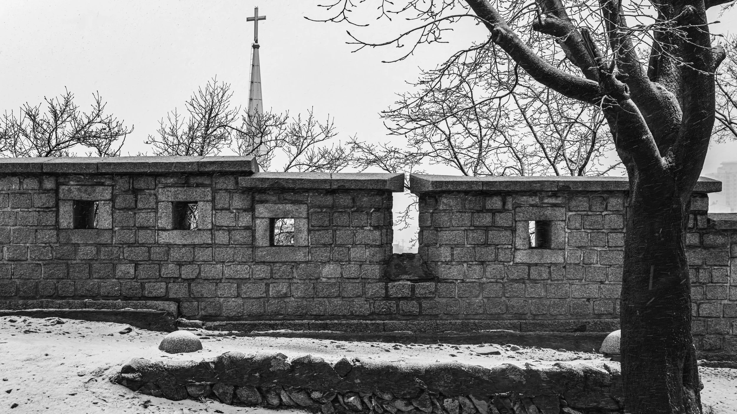 A black and white photo of a stone wall with a cross on a steeple in the background, a large tree with snow on its branches, and a snow-covered ground.