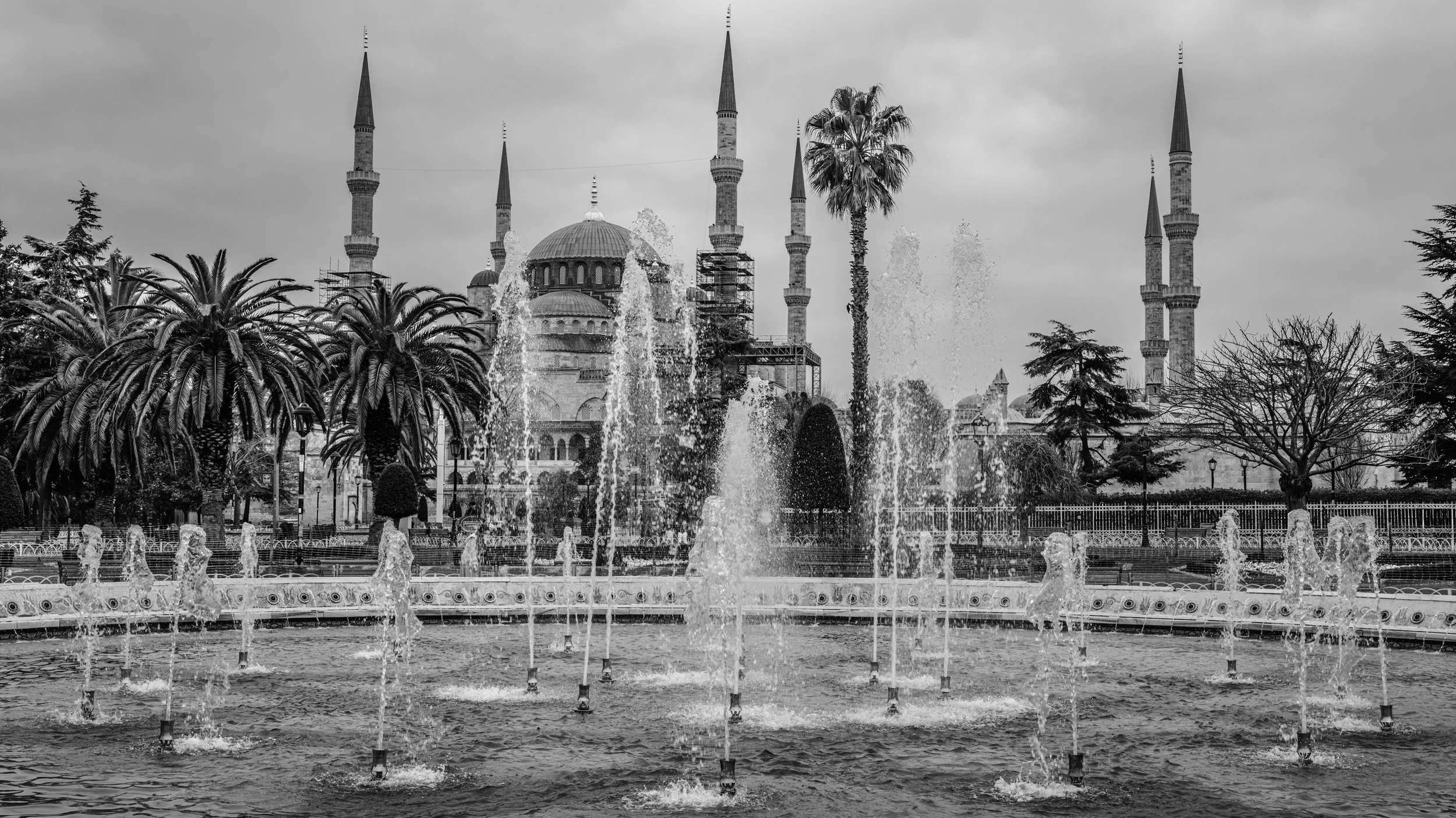 Black and white photo of a mosque with tall minarets, surrounded by palm trees and other trees, with fountains in the foreground.