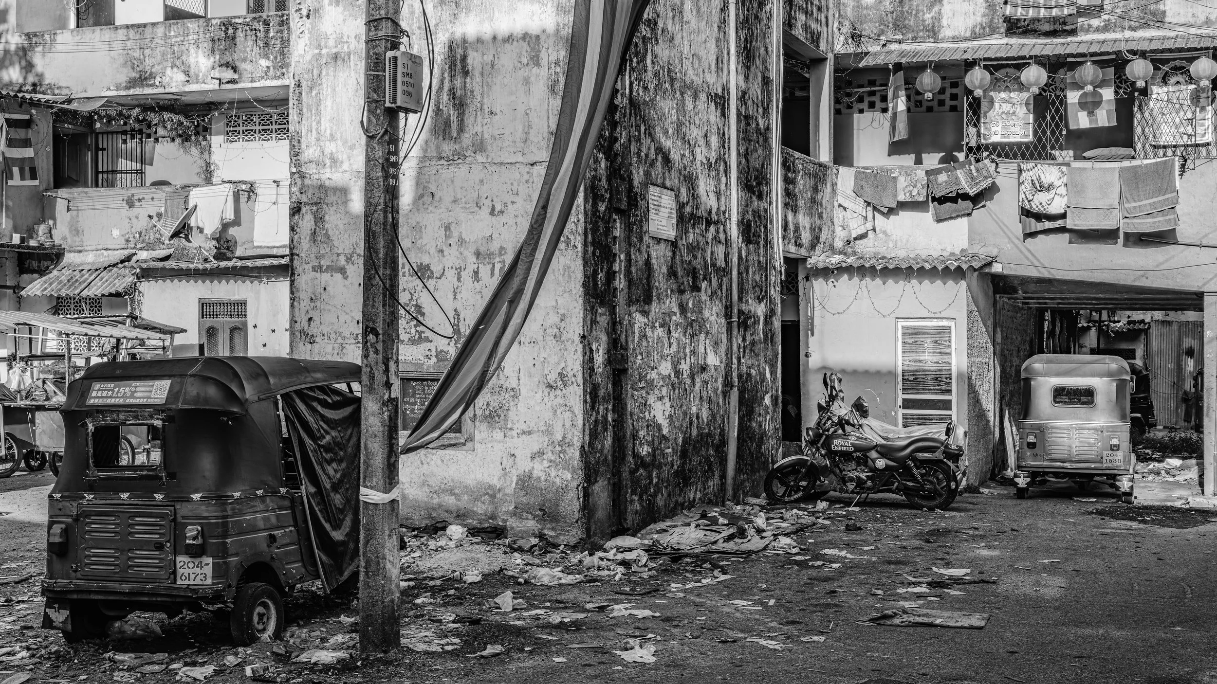 A black-and-white photograph of an urban alley showing a weathered wall with hanging laundry, a motorcycle, an auto rickshaw, and various debris on the ground.