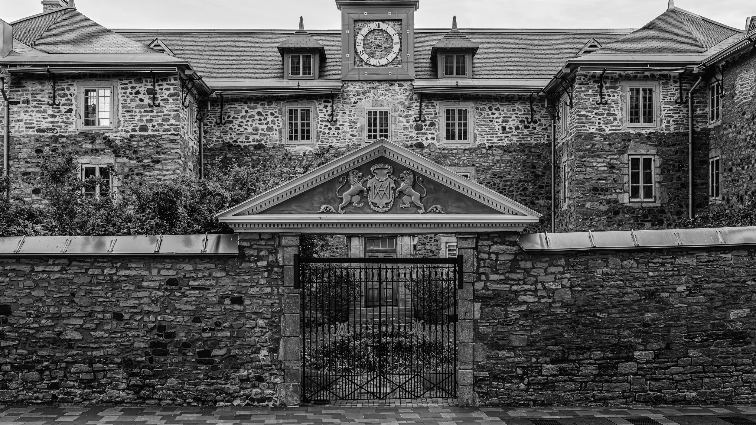 Black and white photo of a stone building with a clock tower, decorative crest with lions, and iron gate in front.