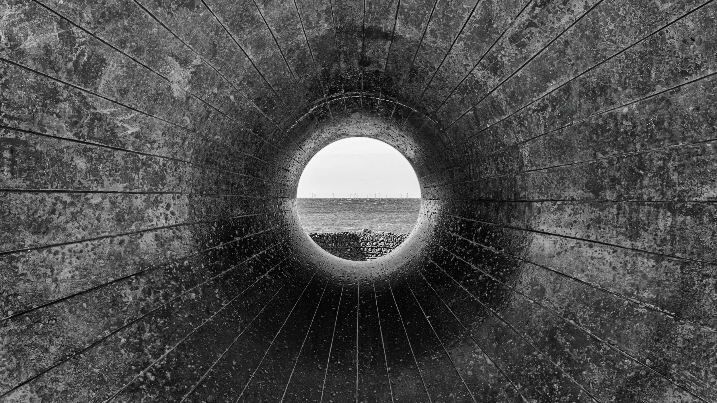 A black and white photo taken through a large, circular, tunnel-like structure with concentric, textured surfaces and thin, evenly spaced lines radiating outward from the center. In the distance beyond the tunnel, there's an open body of water with w