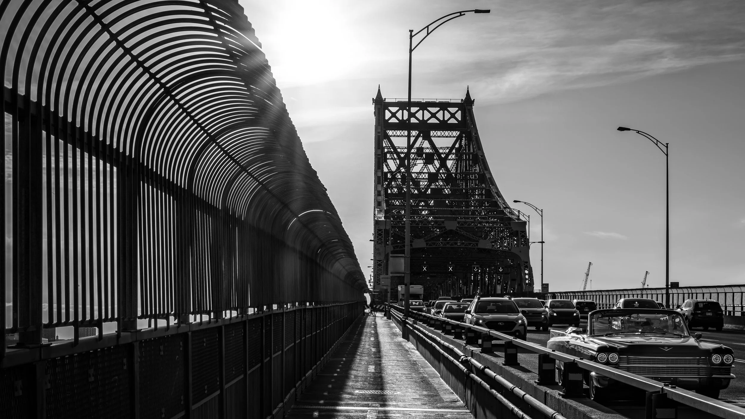 Black and white photo of a bridge with a large structure on the right and a fenced walkway with cars on the left. The sun is behind the bridge, creating a silhouette effect.