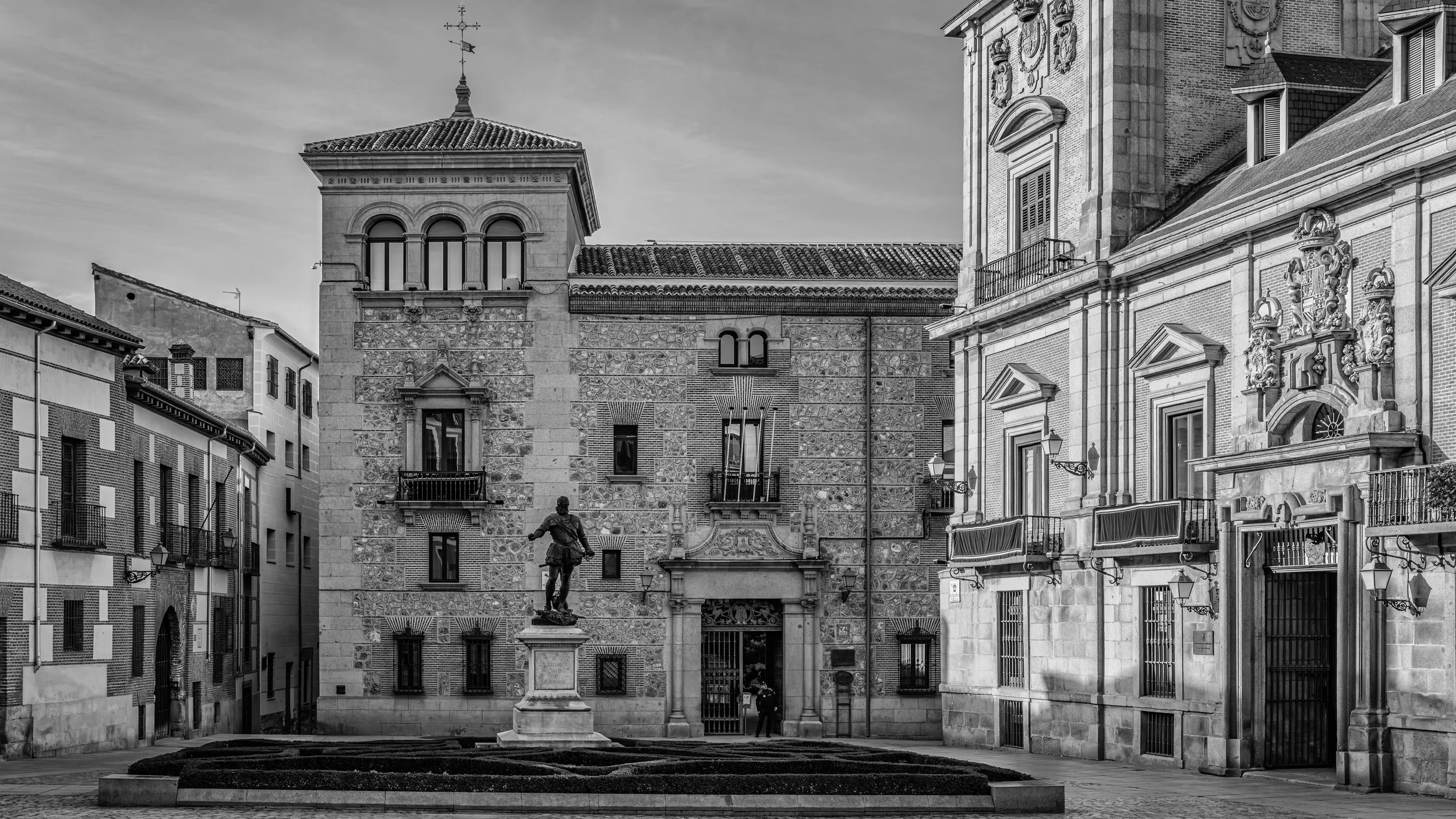 Black and white photo of historic European buildings with ornate architectural details, a statue of a man on a pedestal, and a courtyard with cobblestone pavement.