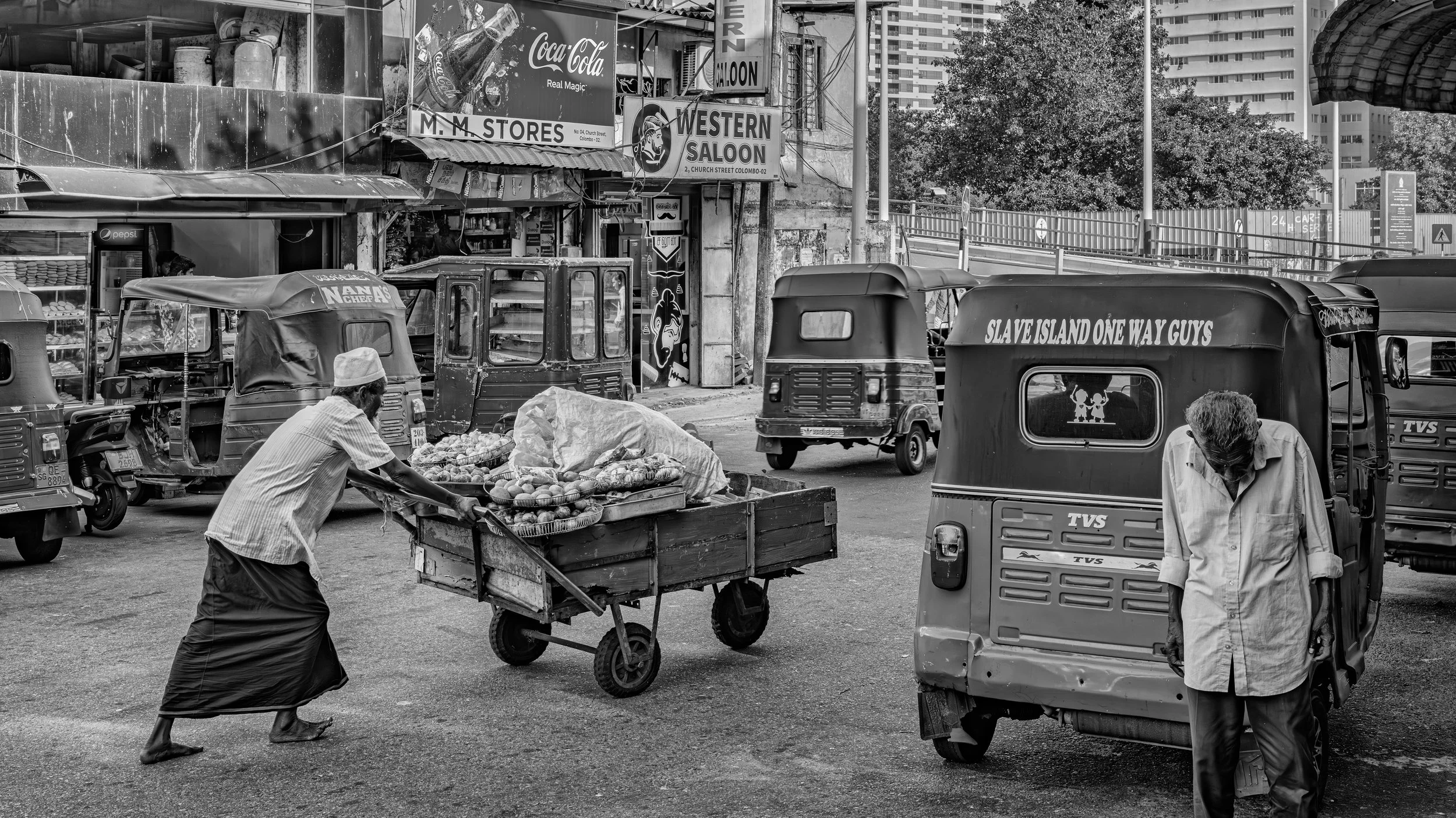 Street scene in an urban area with two women and a man, three auto rickshaws, a cart filled with fruits and vegetables, signage for stores and a saloon, and multi-story buildings in the background.