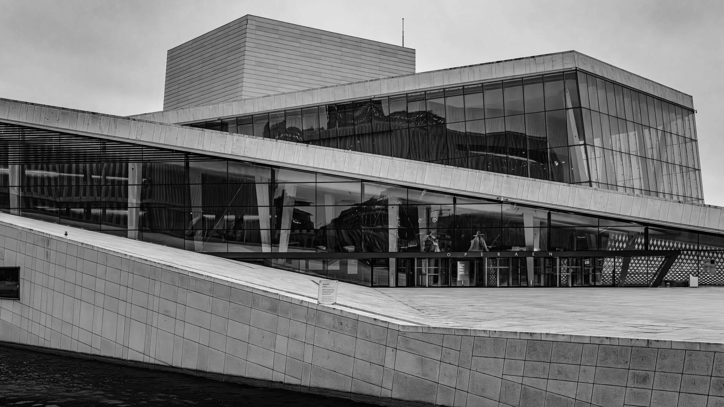 Modern building with glass windows and angular architecture, reflecting mountains and sky, situated along water with a walkway in the foreground.