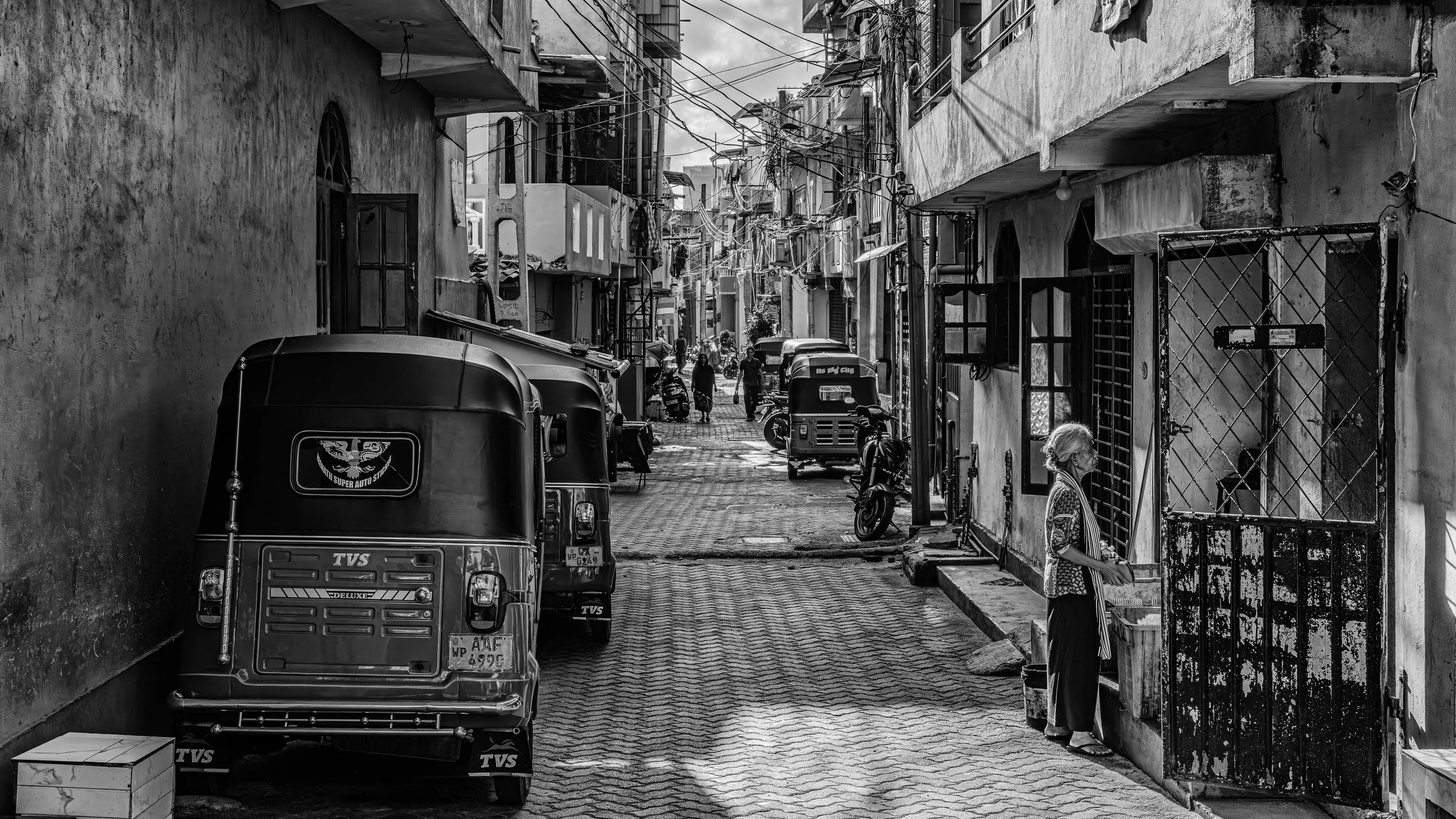 A narrow alleyway in an urban neighborhood with parked vehicles, power lines overhead, and a woman standing near a gated door on the right side.