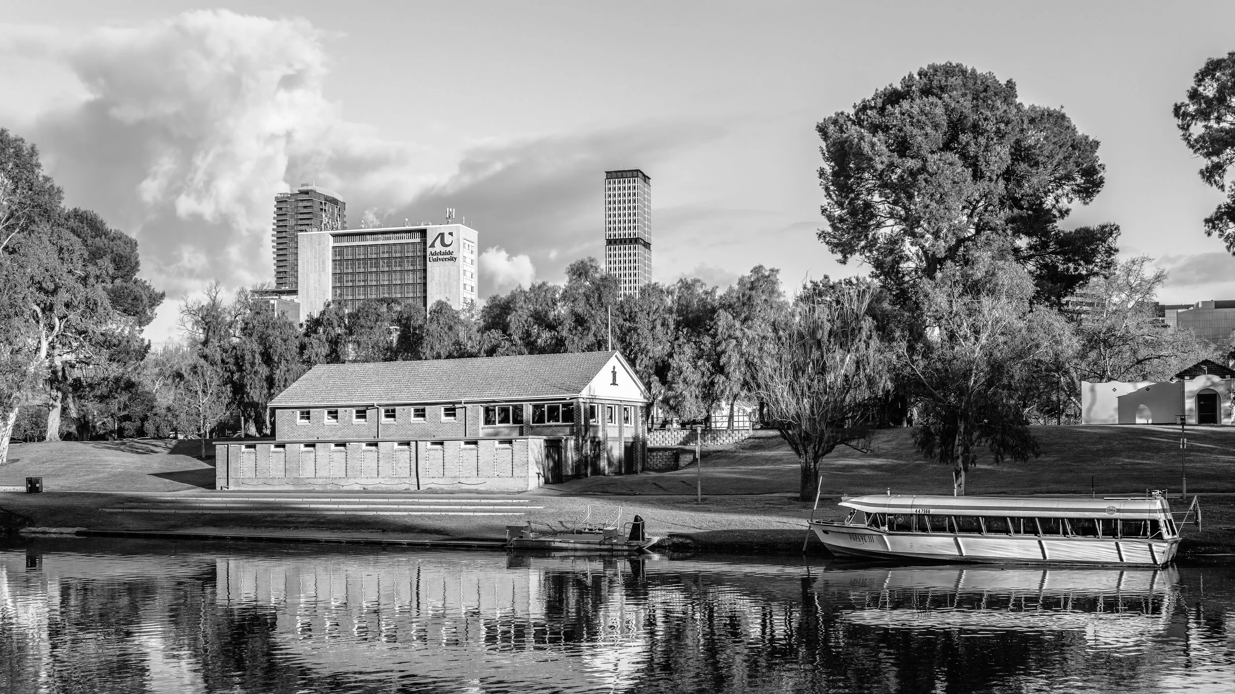 Black and white photo of Adelaide University buildings viewed across a river, with trees in the foreground and a boat on the water. Adelaide, Australia.