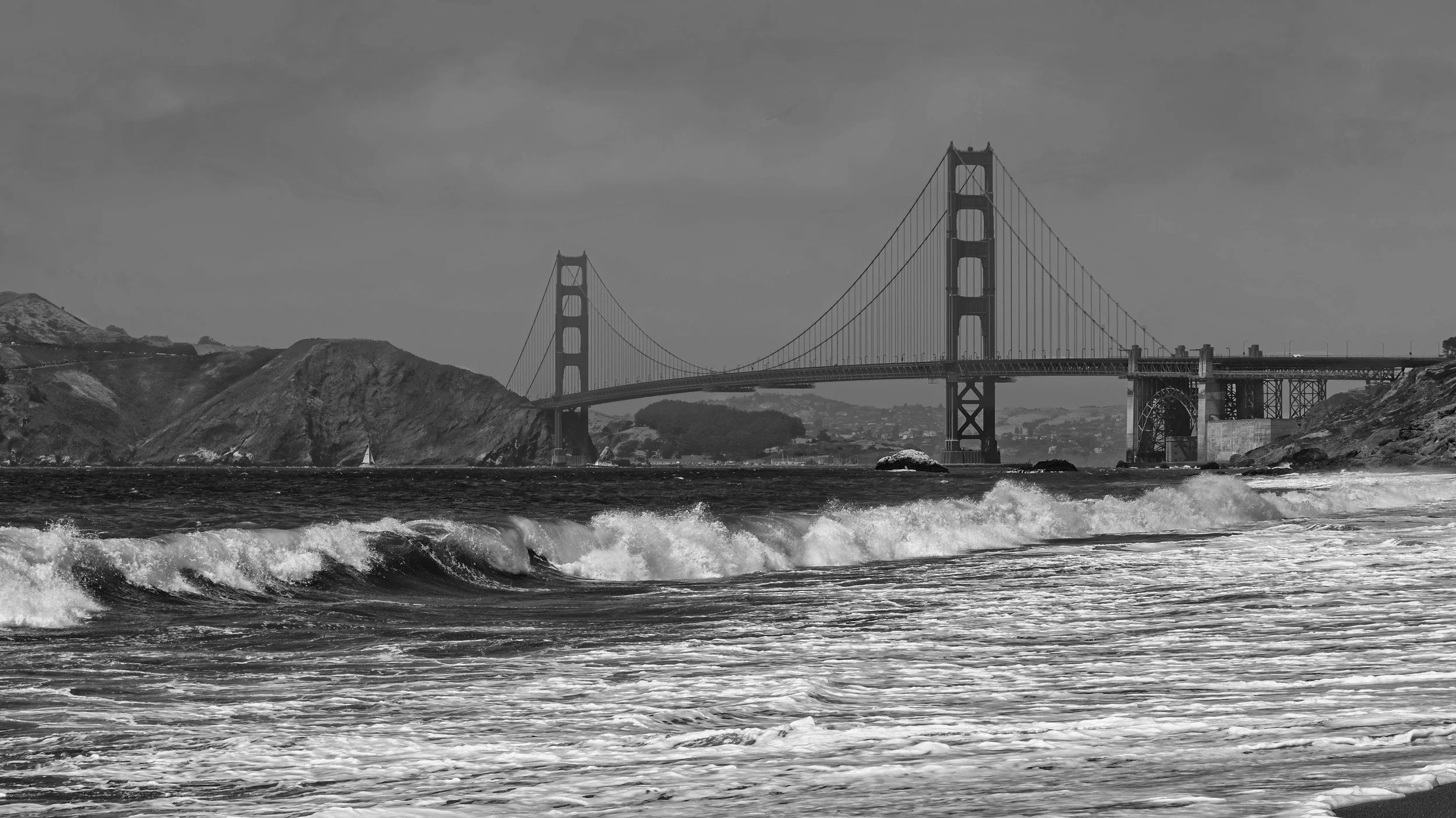 Black and white photograph of the Golden Gate Bridge with water and waves in the foreground and hills in the background.
