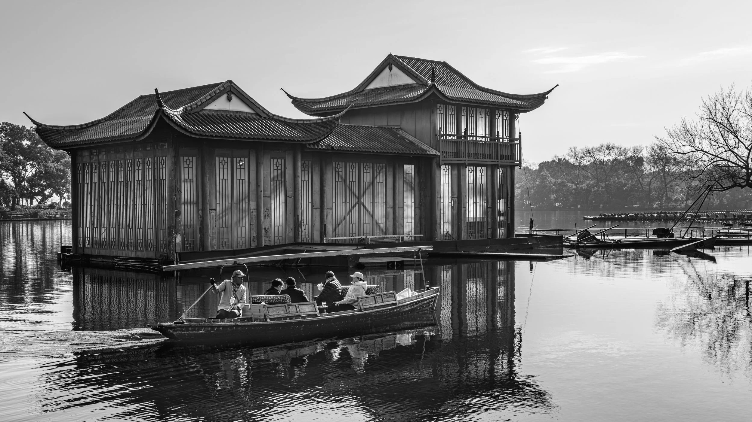 A black and white photo of a waterside traditional Asian-style building with a boat carrying people passing in front of it. Trees and a calm body of water are in the background.