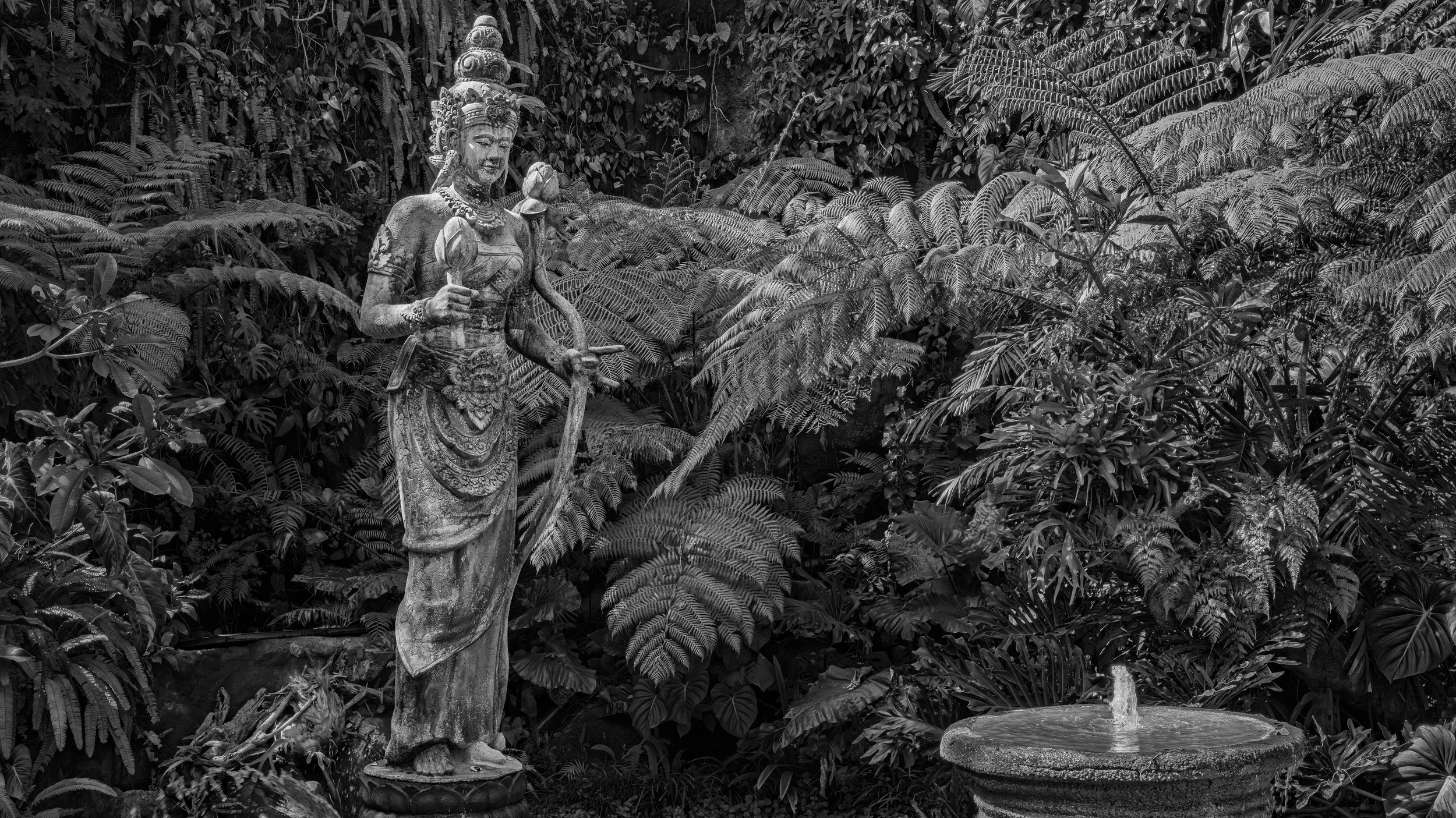A stone statue of a woman holding a staff in a lush, tropical garden with dense ferns and foliage.