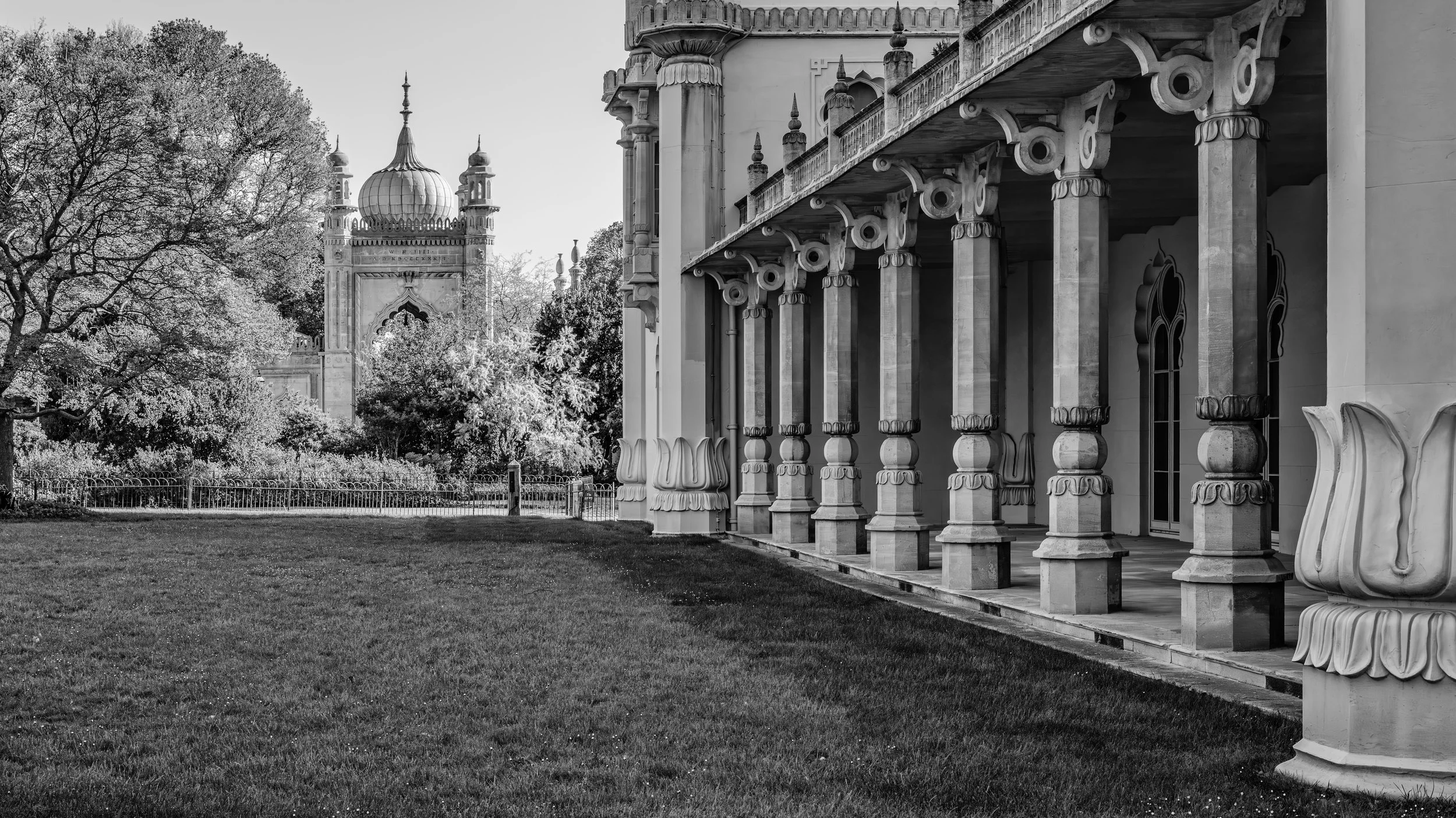 Black and white photo of a historic Indian palace with intricate architectural features like arches and columns, set next to a manicured garden with trees and a fence, with a tall onion-domed tower in the background.