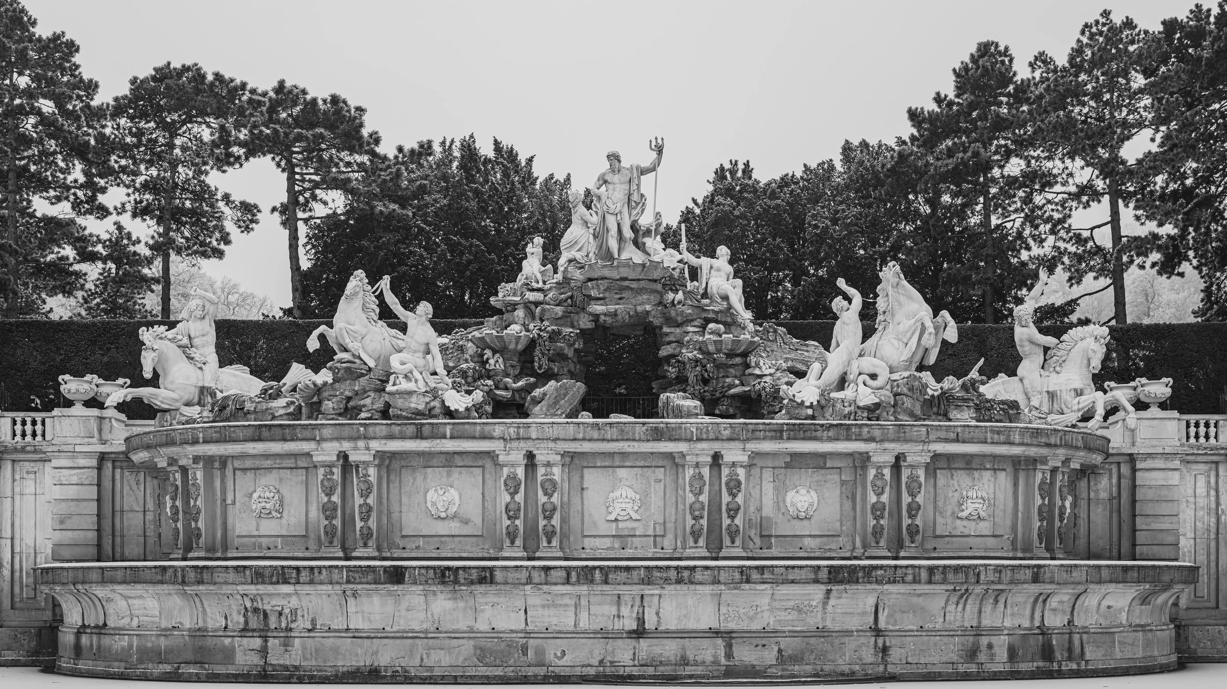 Black and white photograph of a grand detailed fountain with various mythological figures and sculptures, surrounded by trees.