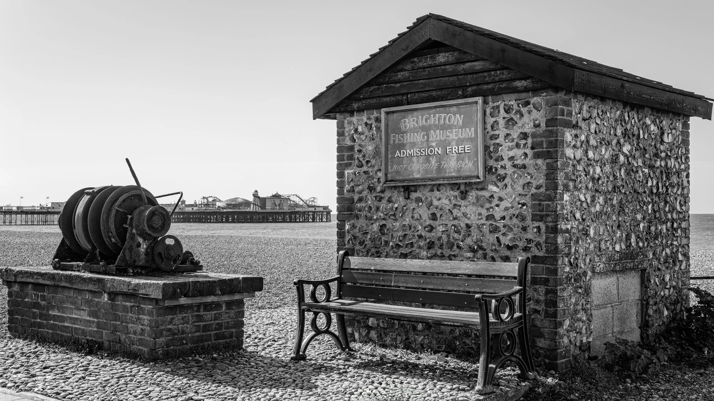 A historic brick and stone structure with a sign for Brighton Fishing Museum, a bench, a vintage winch, and a view of a pier in the background on a pebble beach, black and white photo.