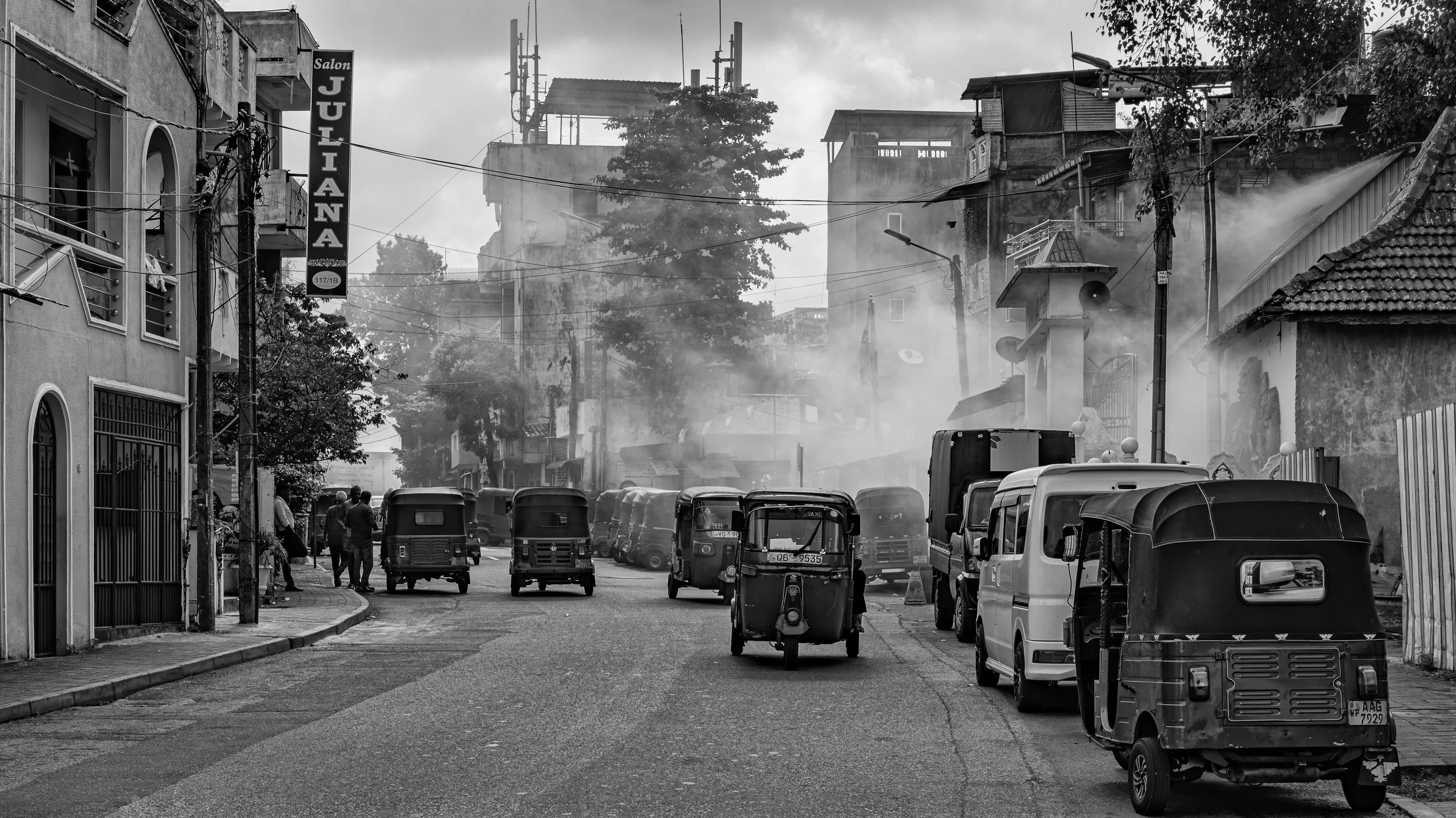 A black and white street scene with multiple auto rickshaws and a few cars parked along the road, some people walking, a sign on a building reads 'Salon Julliana,' and there is smoke rising from behind the buildings, with electrical wires and trees v
