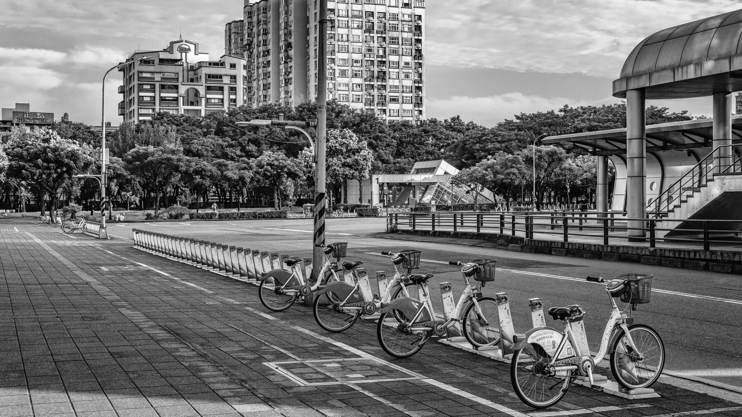 Black and white cityscape with high-rise buildings, trees, bicycles parked on the sidewalk, and a modern bus stop structure.